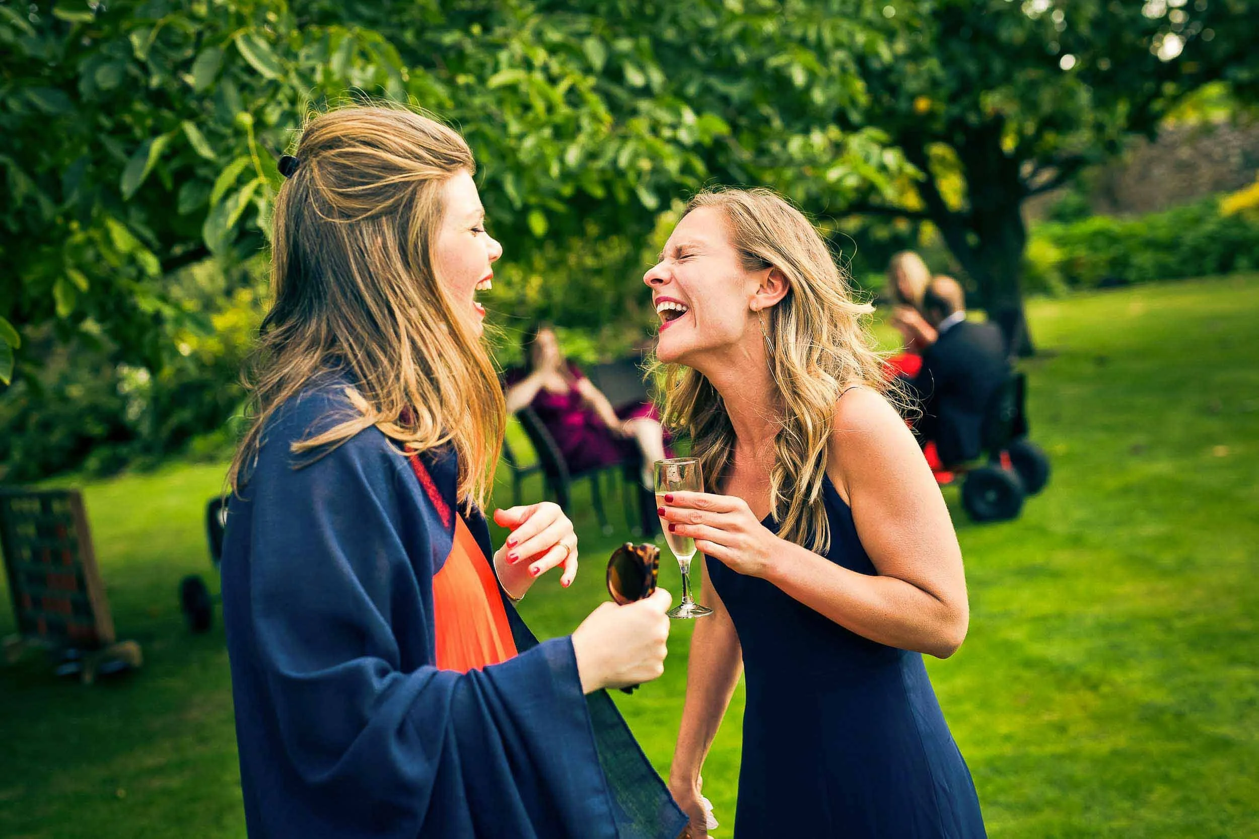 Two women laughing and talking at an outdoor celebration, one holding a glass of champagne and the other holding sunglasses, in a garden with trees and other guests in the background.