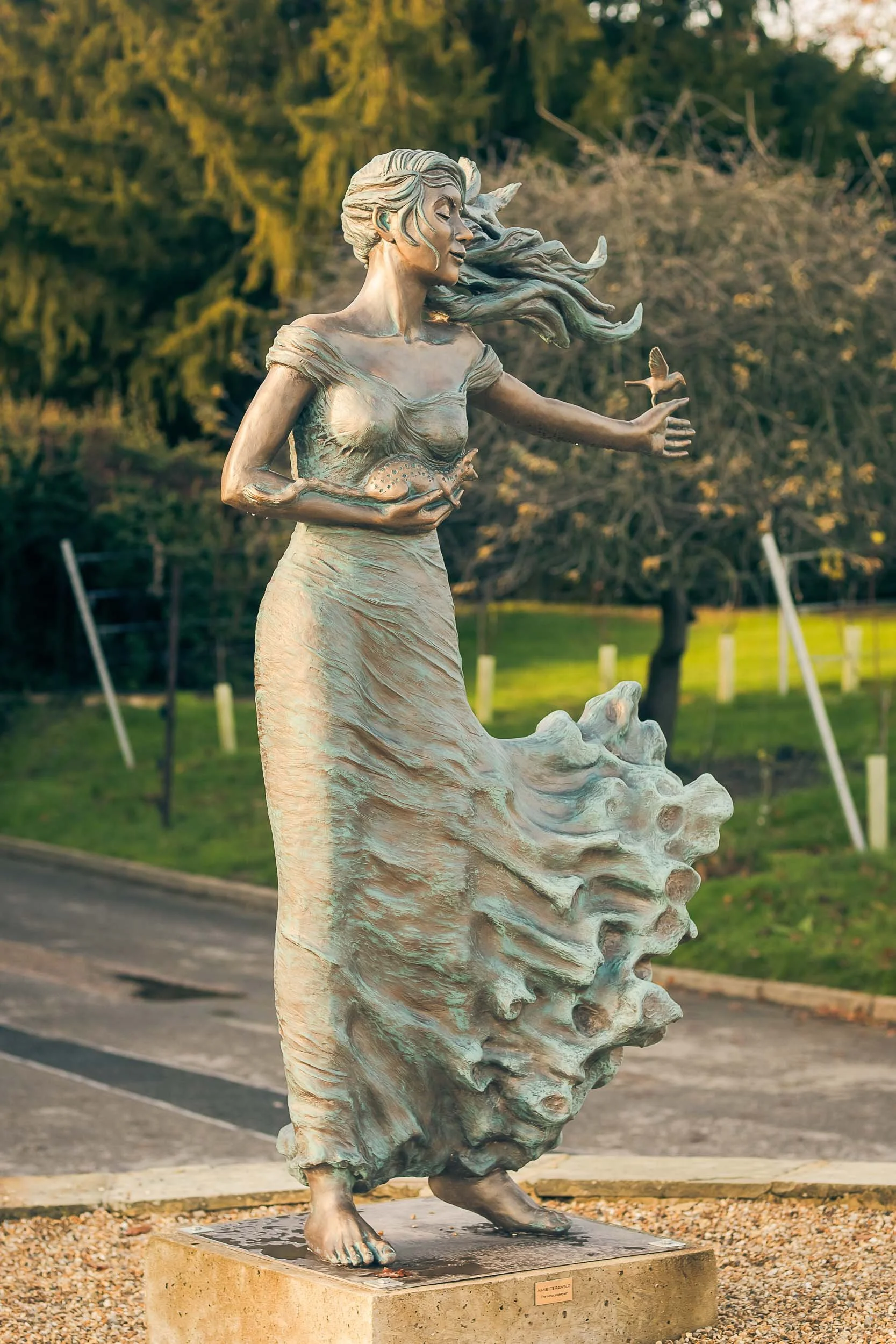 A bronze statue of a woman with long, flowing hair, holding a shell in her left hand and a small bird in her right hand, standing outdoors on a concrete base with trees and a grassy area in the background.