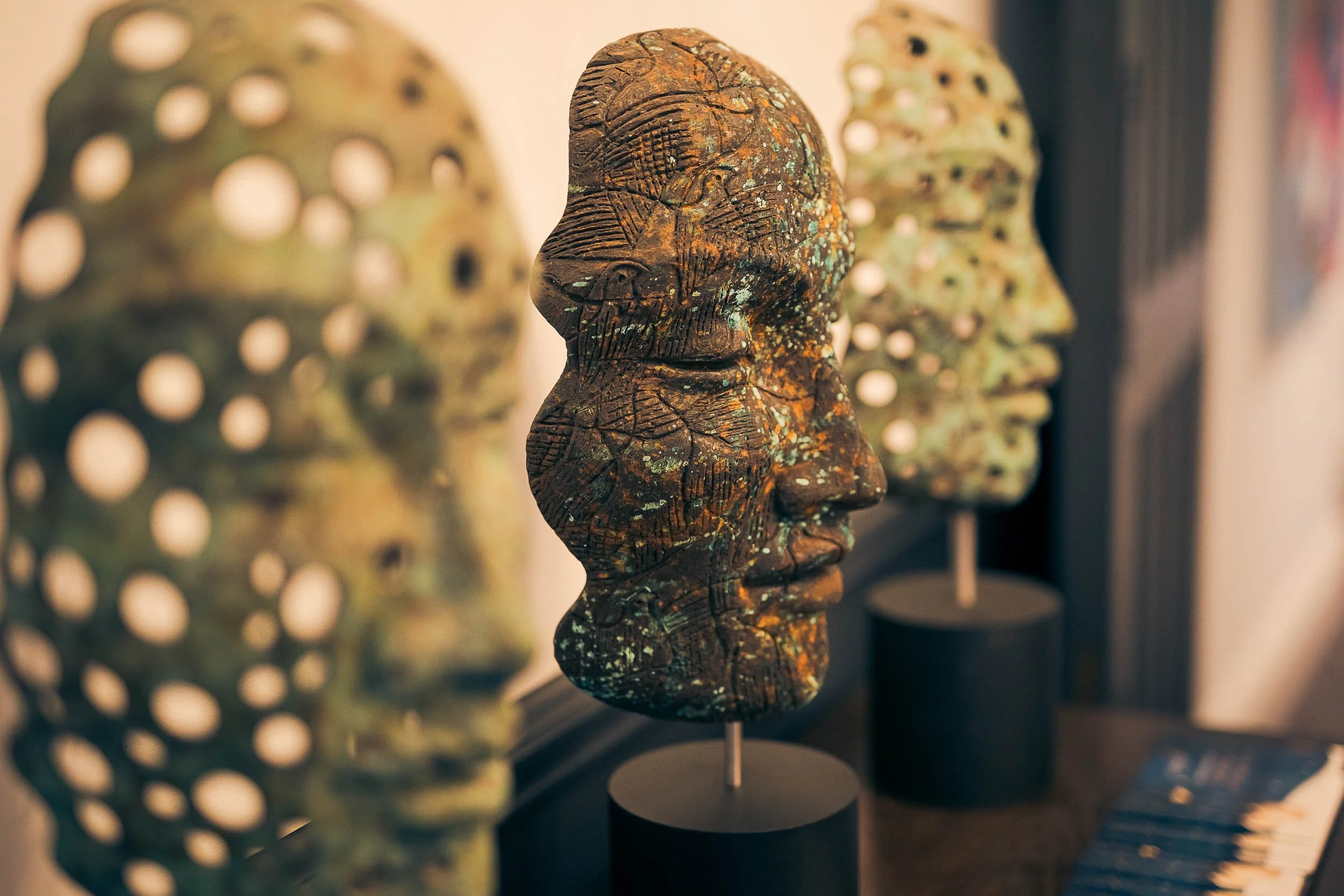 Sculptural art pieces depicting human faces, with textured and patterned surfaces, arranged on black stands in an indoor gallery setting.