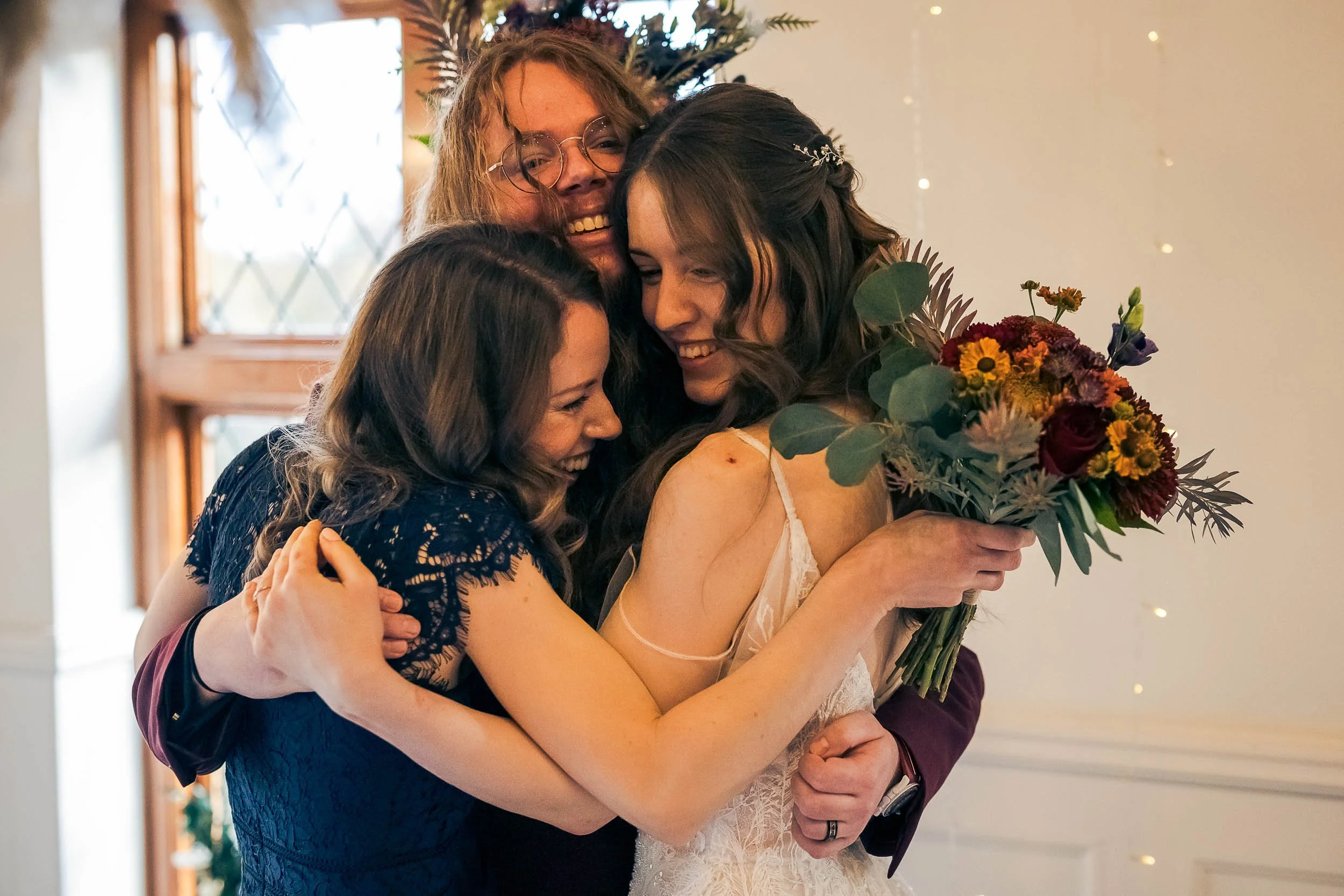 Four women hugging at a celebration, with one woman holding a bouquet of flowers and wearing a beige dress. They are smiling and appear happy.