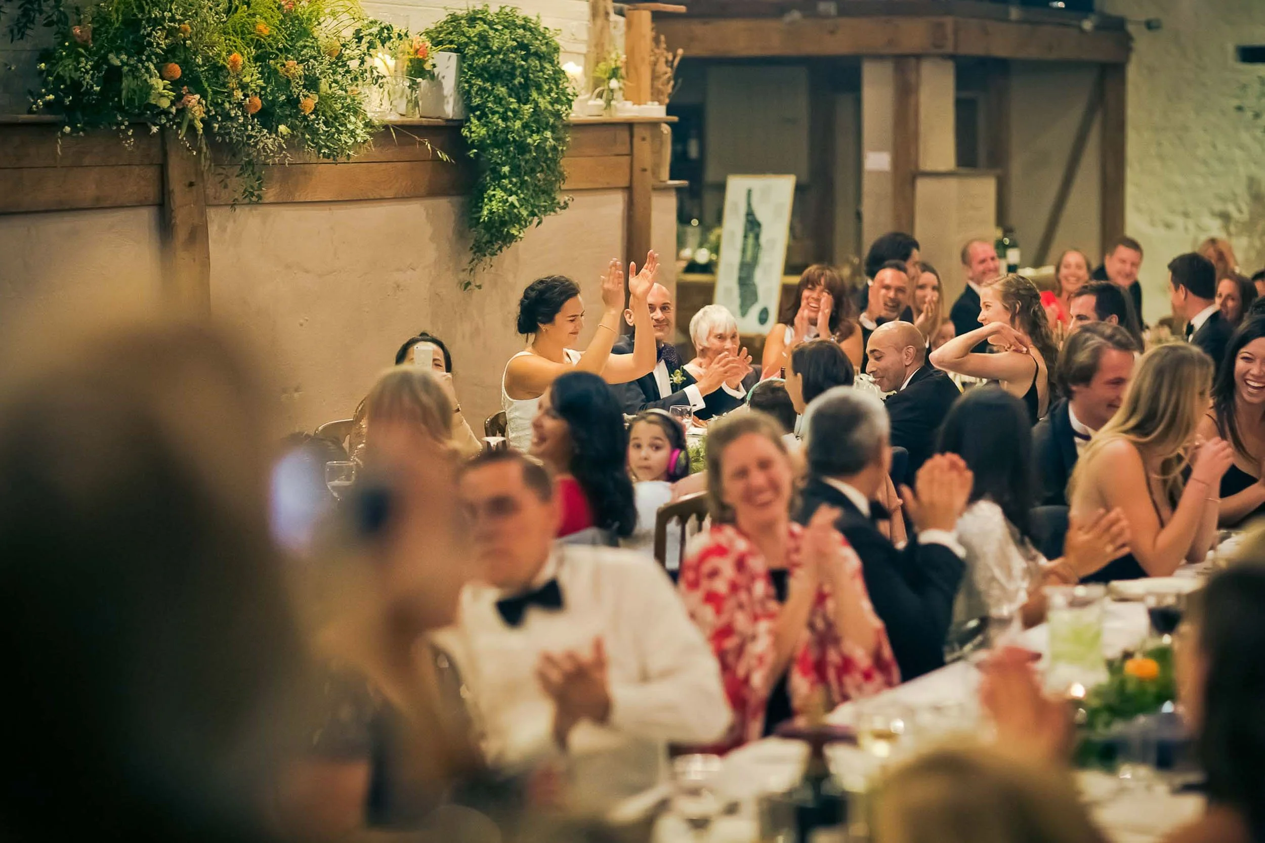 People dressed in formal attire sitting at a long banquet table, applauding and smiling, during a celebration or wedding reception in a rustic indoor venue with wooden beams and decorative plants.
