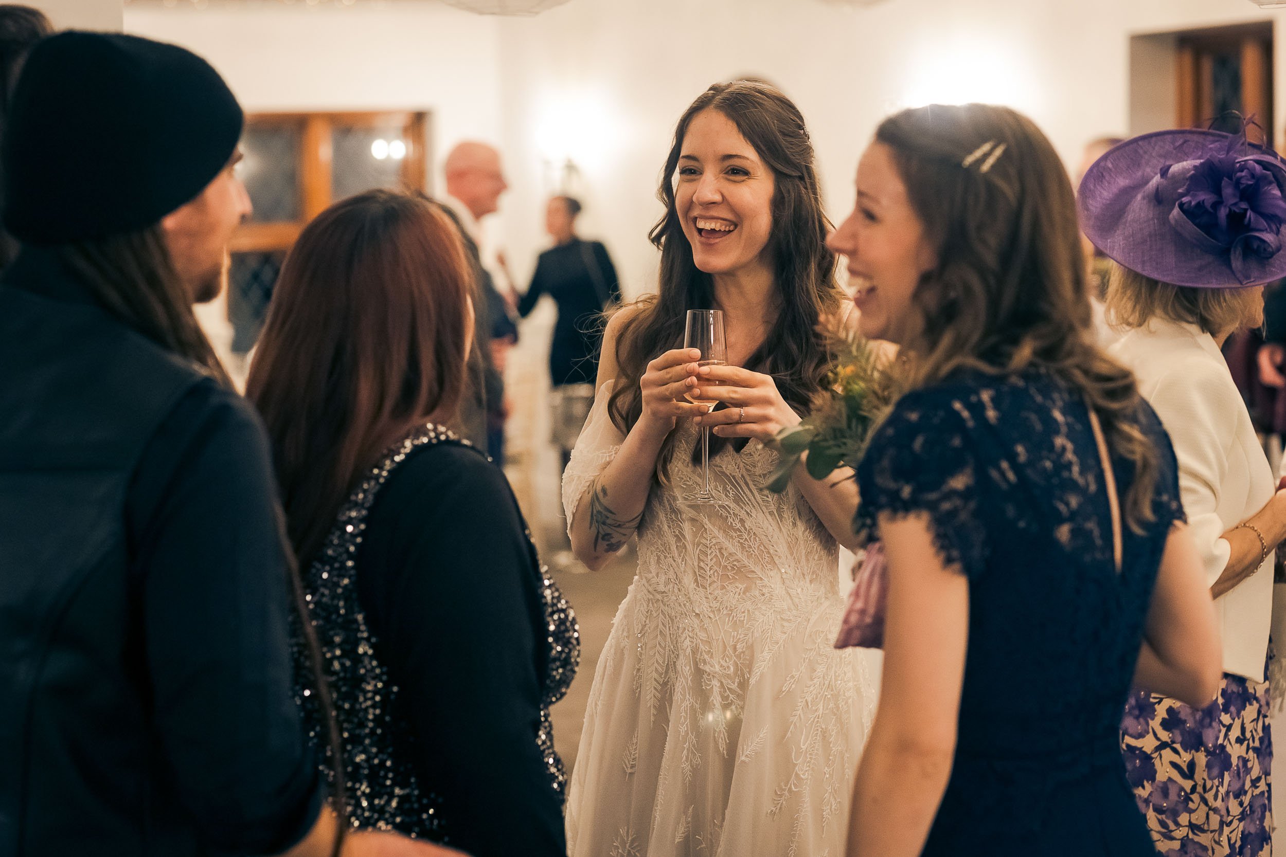 Women in elegant dresses socializing at a wedding or formal event, with a bride holding a champagne flute and smiling.
