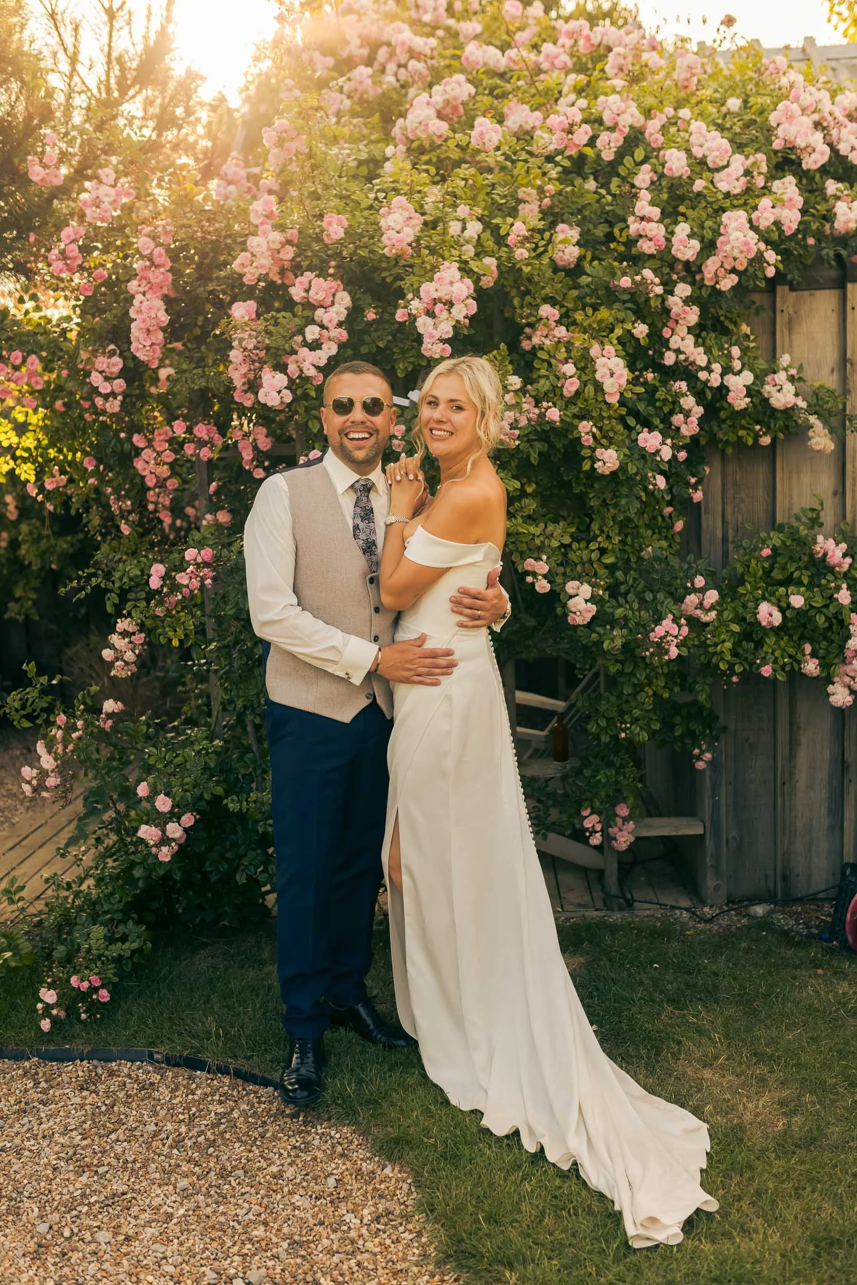 A smiling couple in wedding attire standing outdoors in front of a lush pink flowering bush with sunlight shining through.