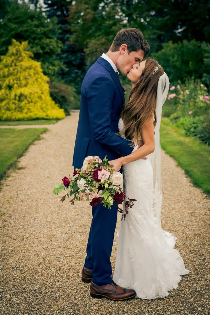 A bride and groom sharing a kiss outdoors on a garden pathway, with the groom in a blue suit and the bride in a white wedding dress holding a bouquet of flowers.