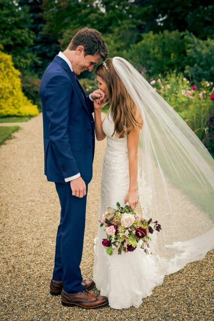 A bride and groom sharing a tender moment outdoors on their wedding day, with the bride holding a bouquet of flowers and wearing a white wedding dress with a veil, and the groom in a blue suit and brown shoes. They are touching foreheads and smiling.