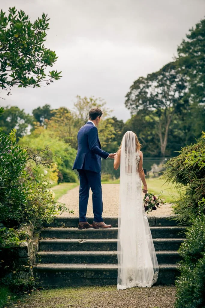 A bride and groom holding hands on outdoor stairs during a wedding, surrounded by green trees and cloudy sky.