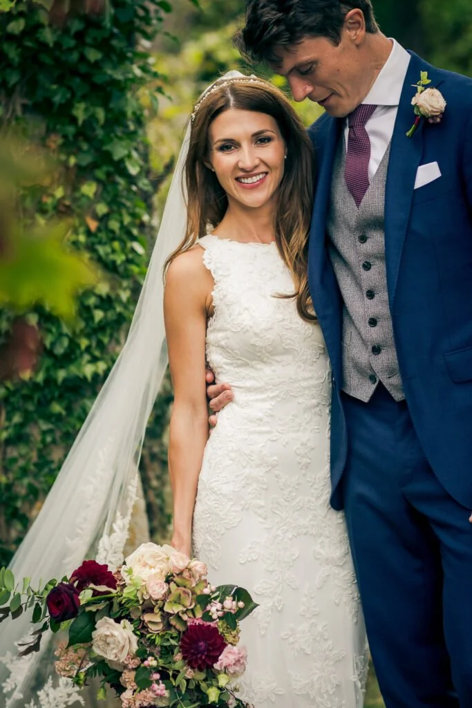 A bride and groom embrace outdoors, smiling, with the bride holding a bouquet of pink, red, and cream flowers and the groom in a blue suit and gray vest.