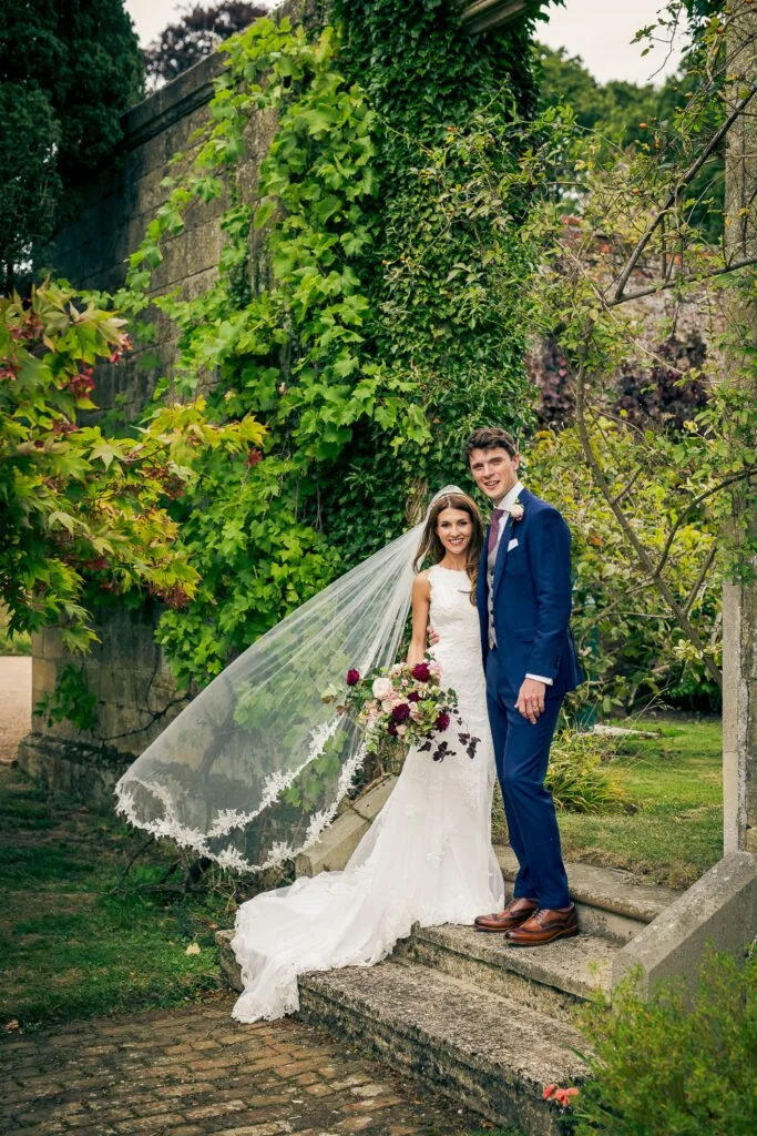 A bride and groom stand together outdoors on a stone staircase, surrounded by lush green foliage. The bride wears a white wedding gown with a long veil, and holds a bouquet of dark red, white, and purple flowers. The groom is dressed in a blue suit w
