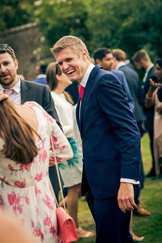 A man in a blue suit smiling and leaning forward, talking to a woman with long hair and a floral dress, at an outdoor event with other well-dressed people in the background.