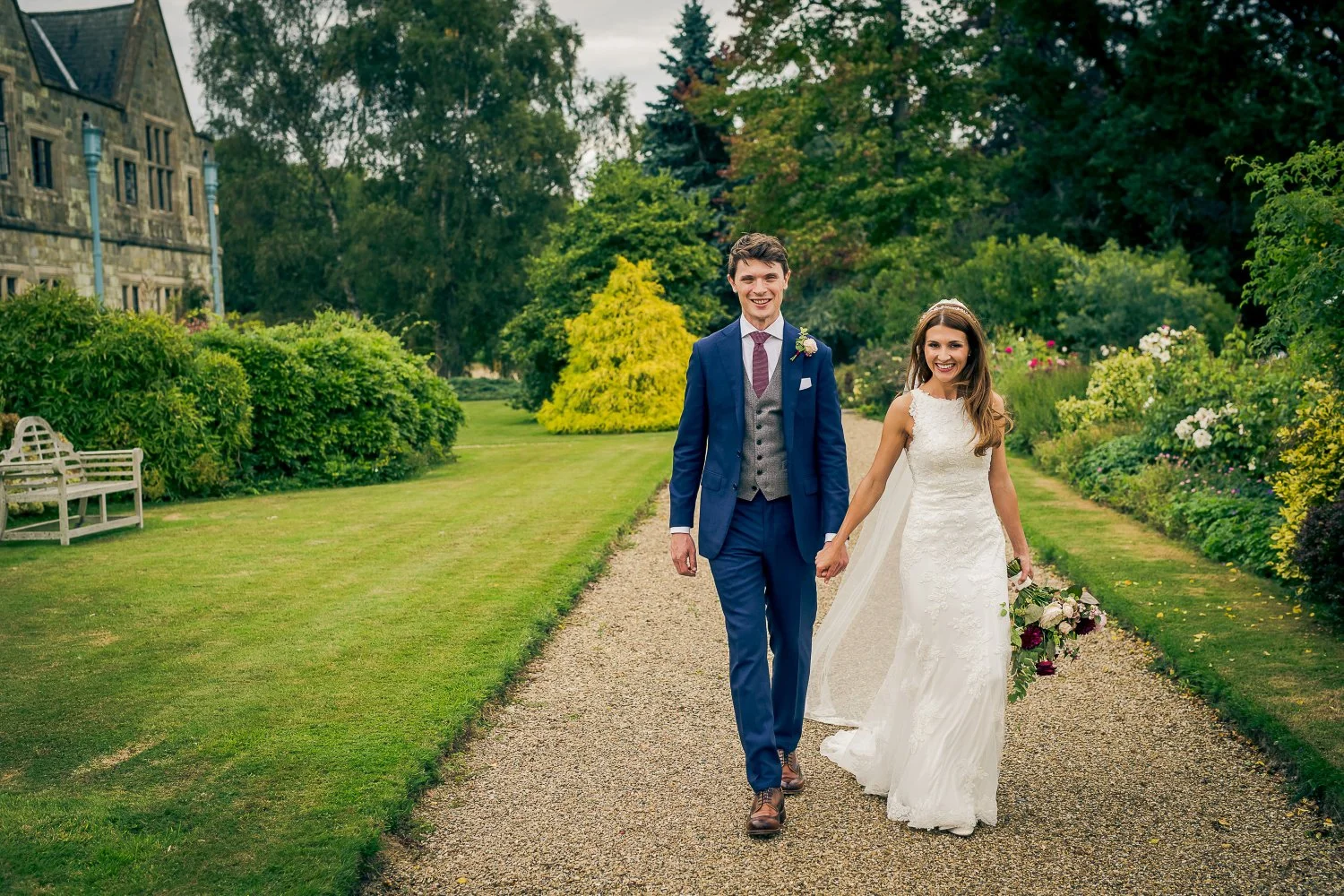 A newlywed couple walking hand-in-hand along a garden path on their wedding day, surrounded by lush greenery and colorful flowers.