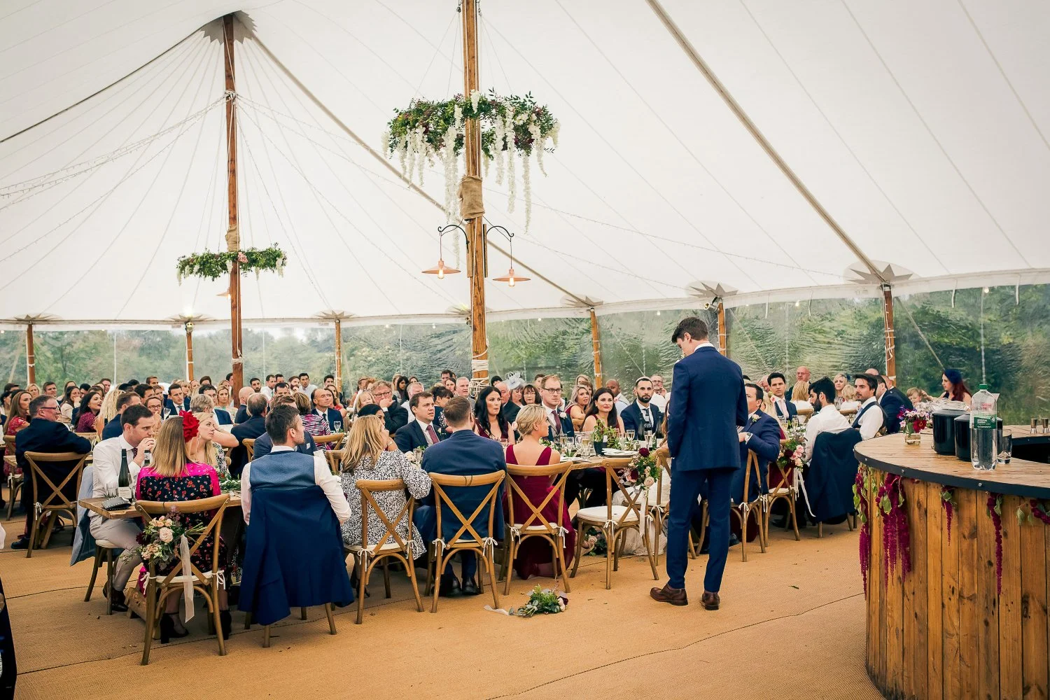 A wedding reception inside a large tent with guests seated at round tables decorated with floral centerpieces, and a man in a blue suit standing. The tent has a white canopy with wooden support poles decorated with hanging greenery and flowers.