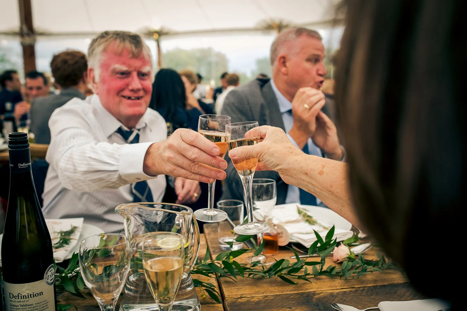 People at a celebration clinking glasses of champagne at a decorated dinner table with wine, glasses of water, and greenery, under a large white tent.