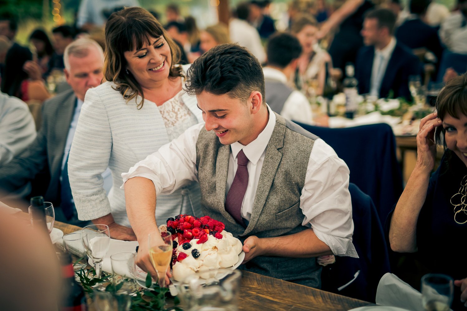 A man in a gray vest and white shirt slices a cake topped with berries during a celebration, with a woman standing beside him smiling. The setting appears to be a lively social gathering or party.