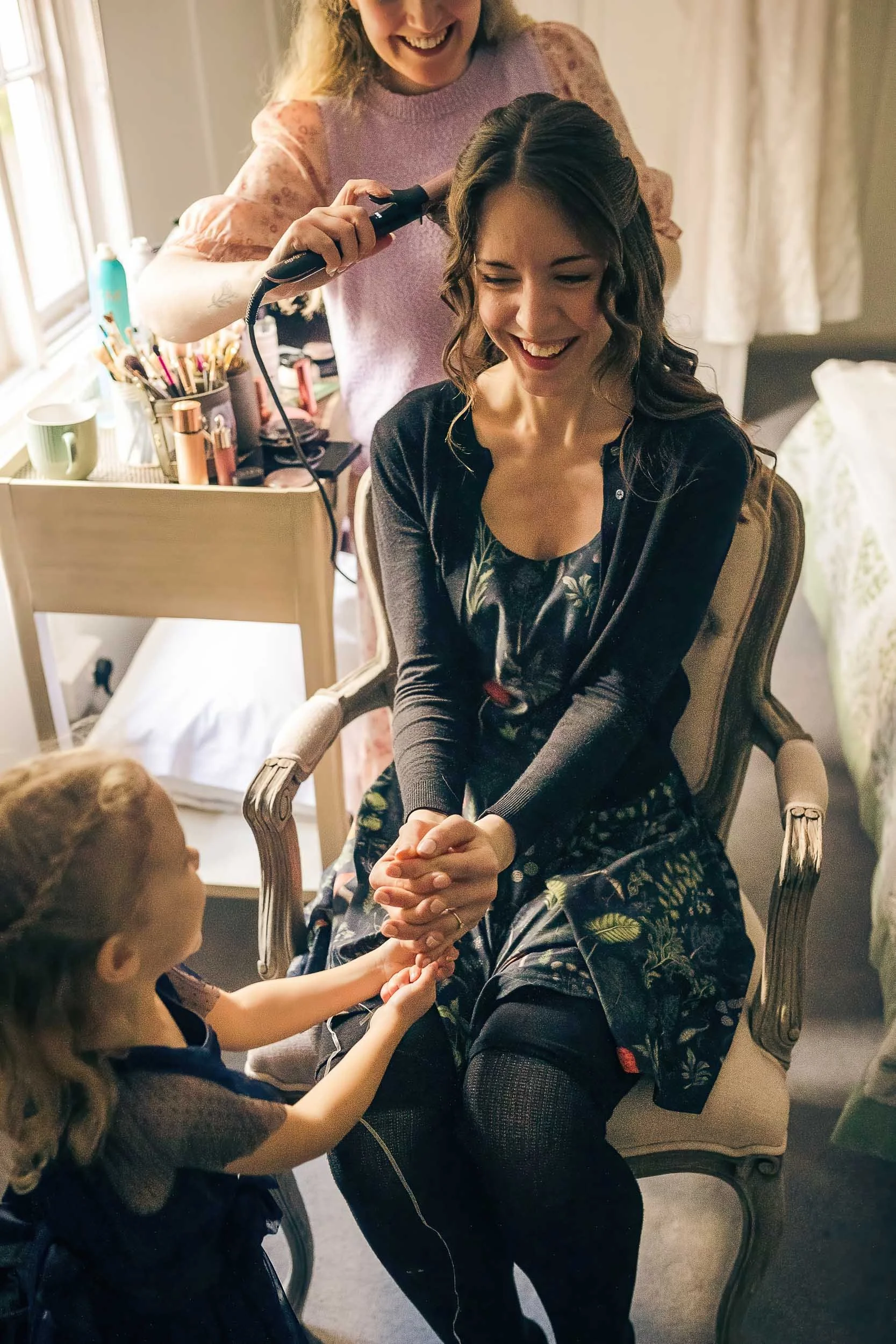 A woman sitting in a vintage chair smiling as she has her hair styled by a hairstylist, while a young girl holding her hands sits in front of her in a cozy room with natural light.