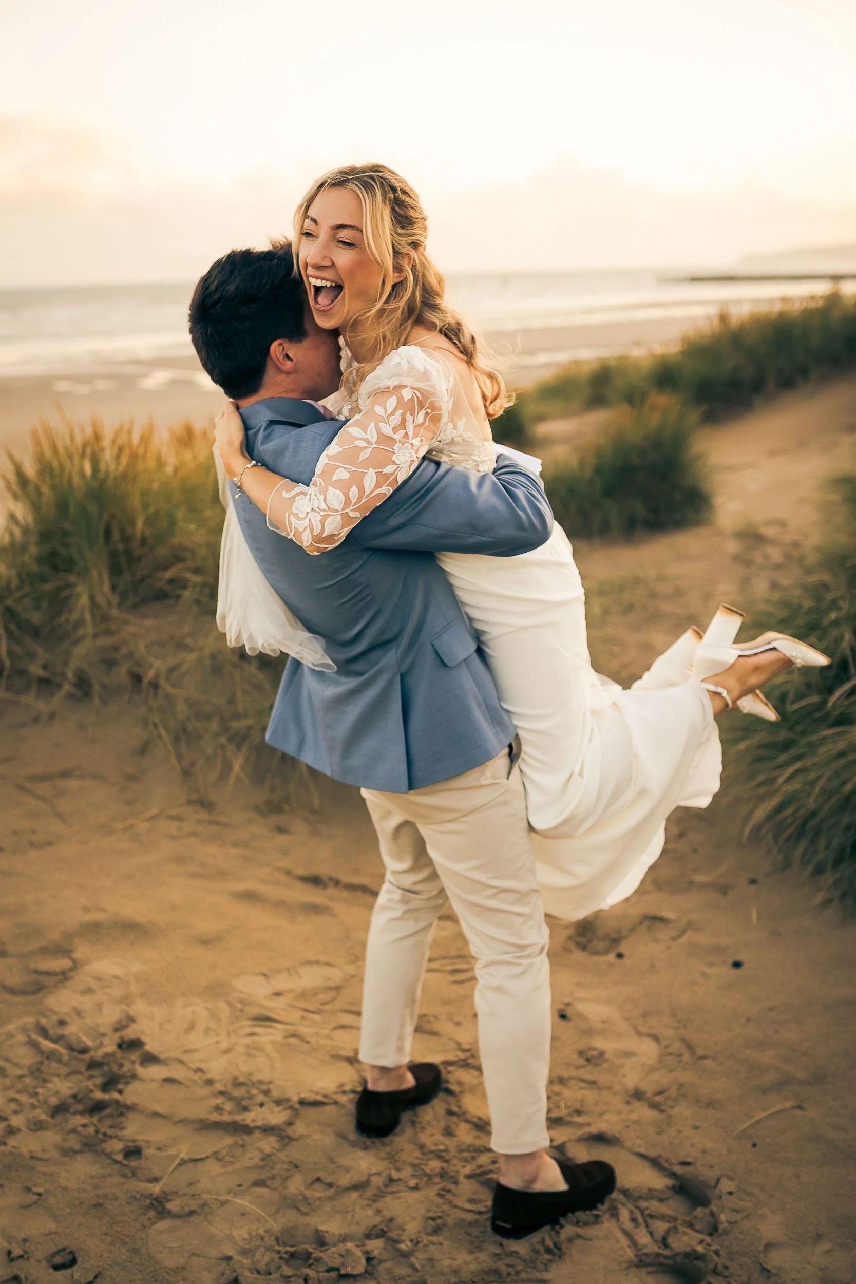 A happy couple on the beach, with the man lifting the woman in his arms as she laughs and smiles, during sunset.