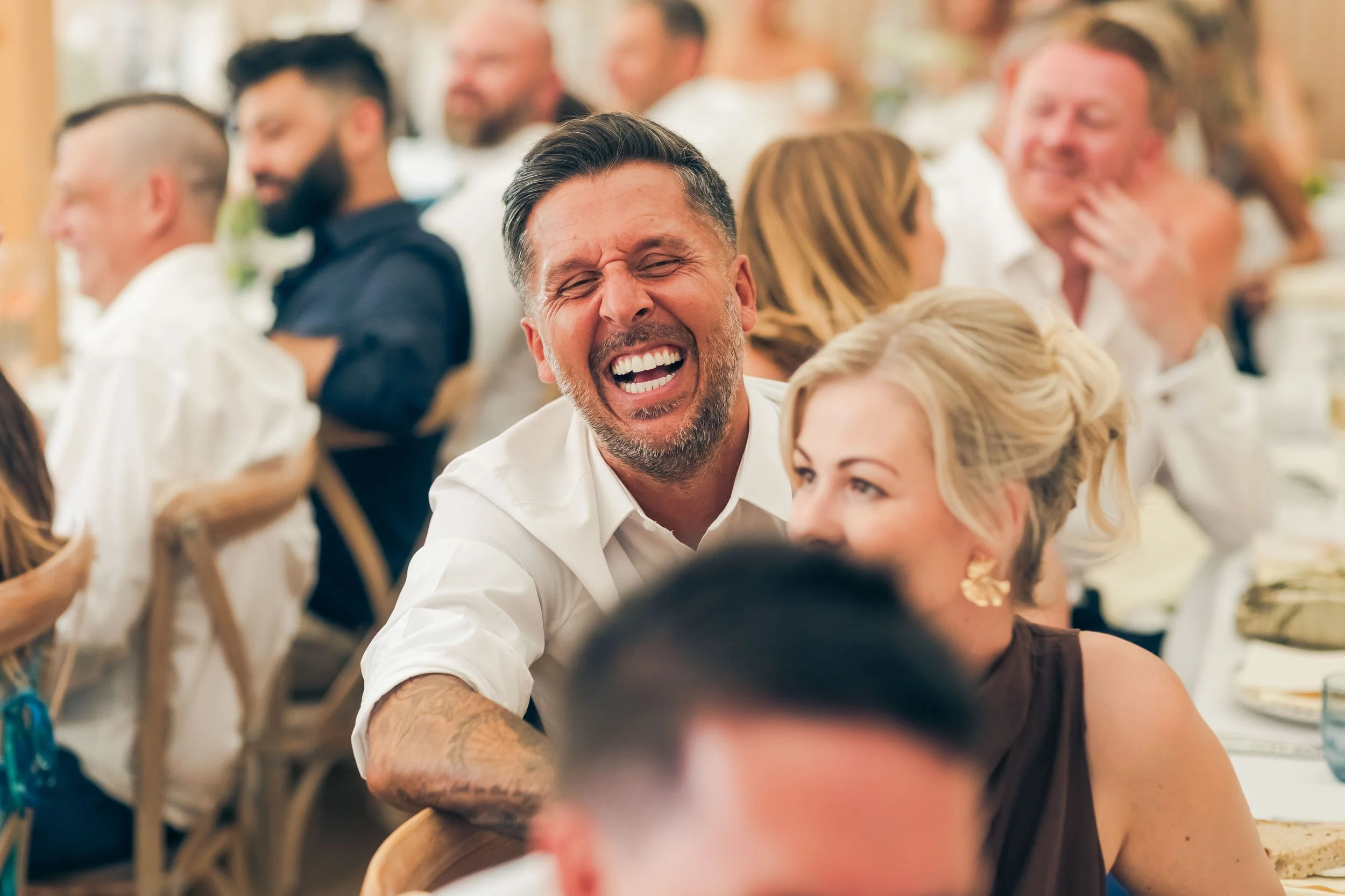 A man with a beard laughing joyfully at a social gathering with other smiling and laughing people, seated at tables in a bright, festive setting.