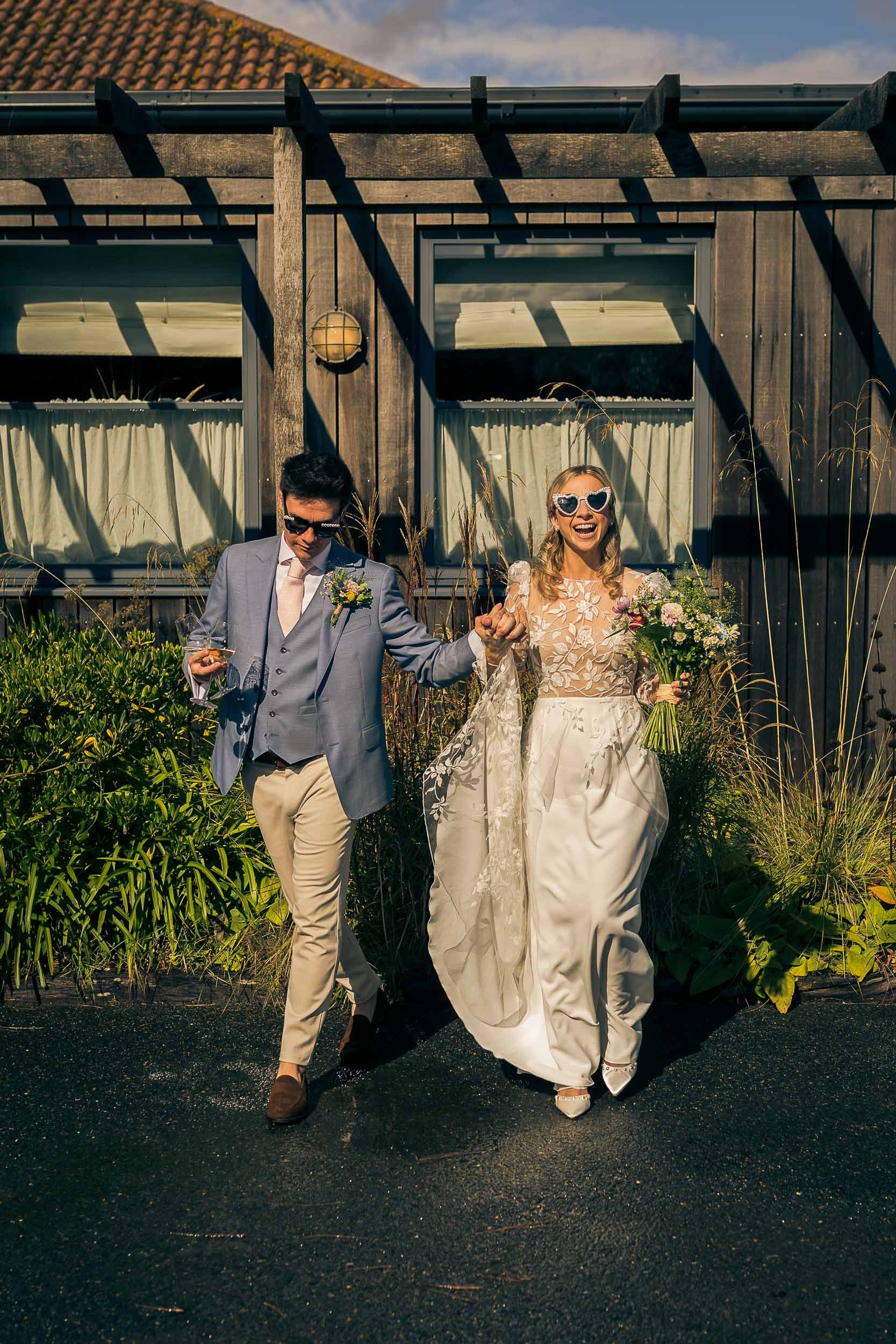 A newlywed couple walking outdoors on a sunny day, holding hands and smiling. The bride is wearing sunglasses, a floral top with a lace overlay, and a long white skirt, carrying a bouquet of flowers. The groom is dressed in a light gray blazer, beige