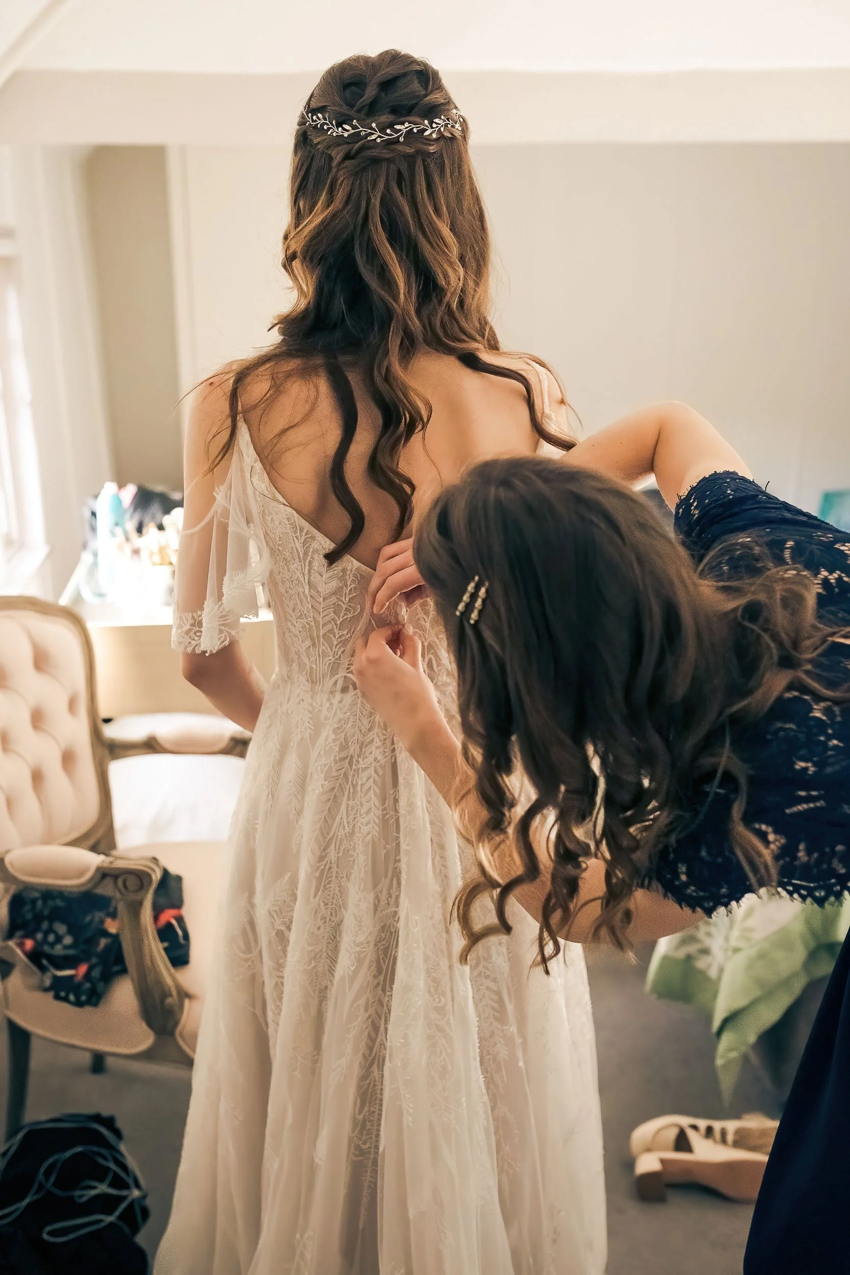 A woman wearing a white wedding dress is having the back buttoned up by another woman in a black lace dress, in a softly lit bedroom with a vintage armchair and a vanity table.