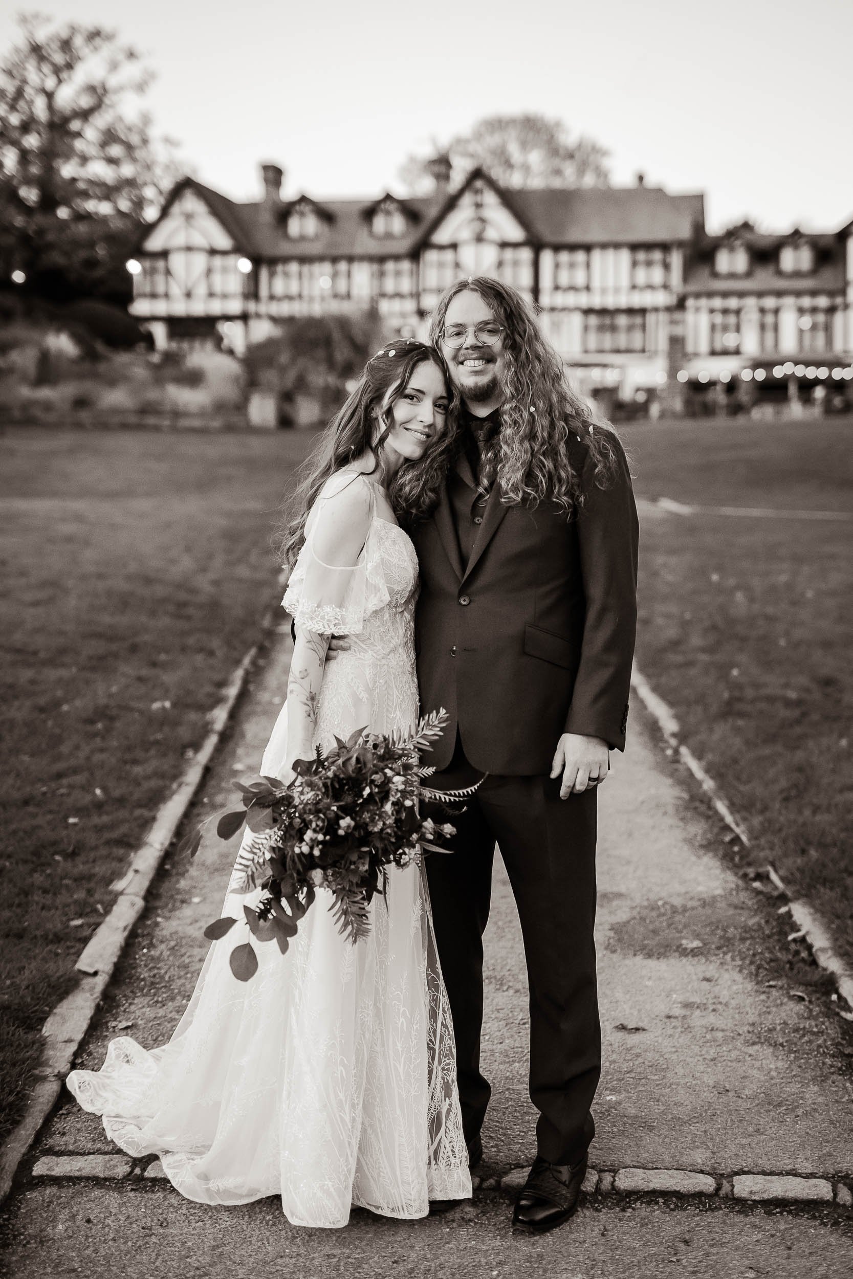 Black and white photo of a bride and groom standing on a walkway outdoors, with a large house in the background. The bride wears a lace wedding dress and holds a bouquet, while the groom wears a suit. They are smiling and hugging.