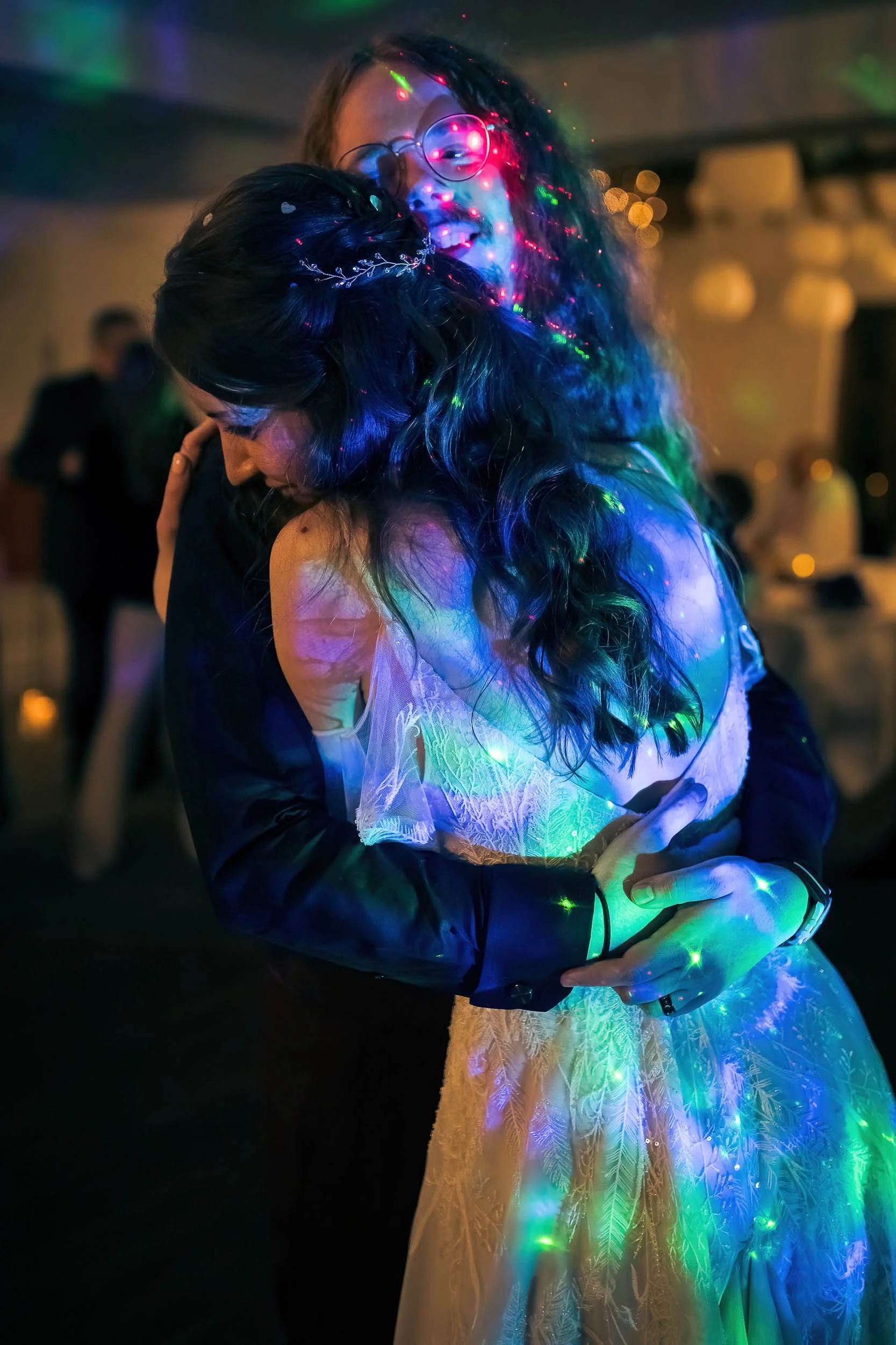 Two people hugging at a celebration or party, with colorful lights projected onto them.