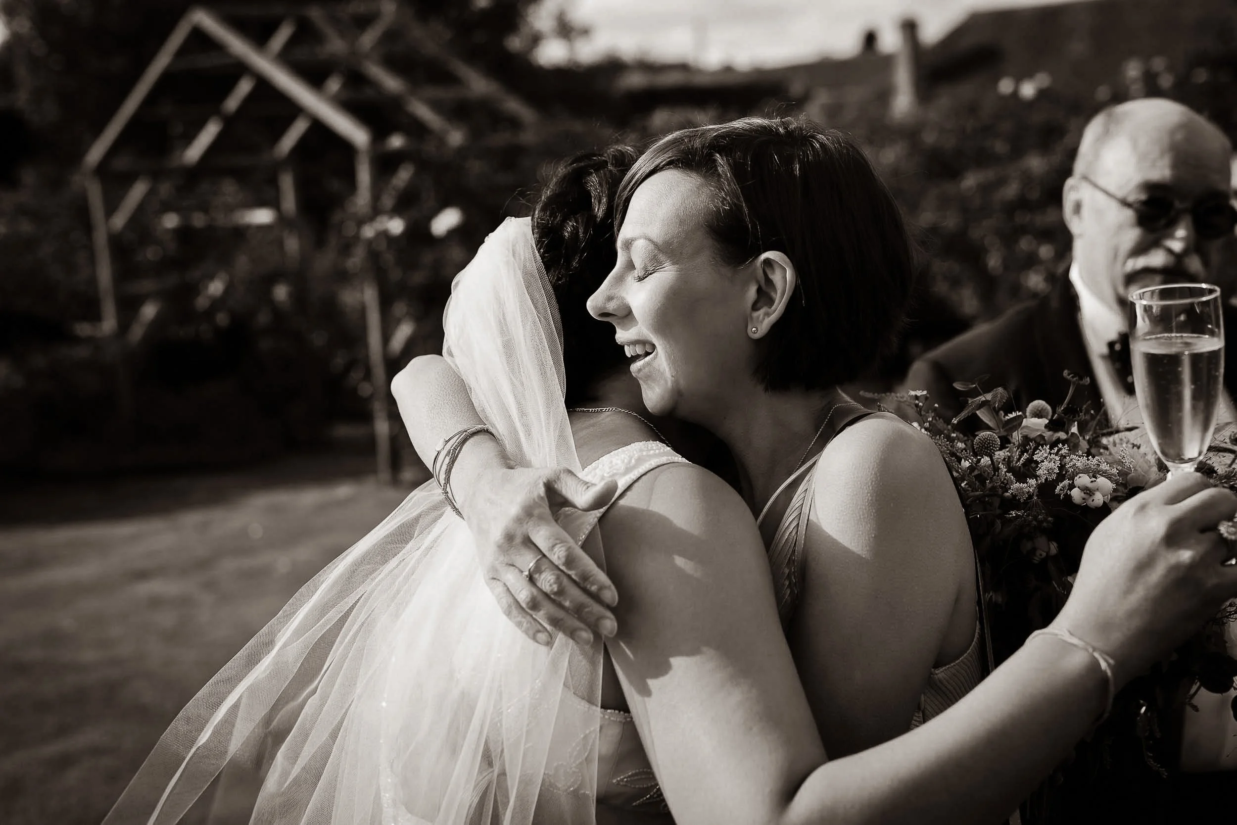 A woman in a wedding dress hugging another woman, both smiling with eyes closed, outside during daytime.