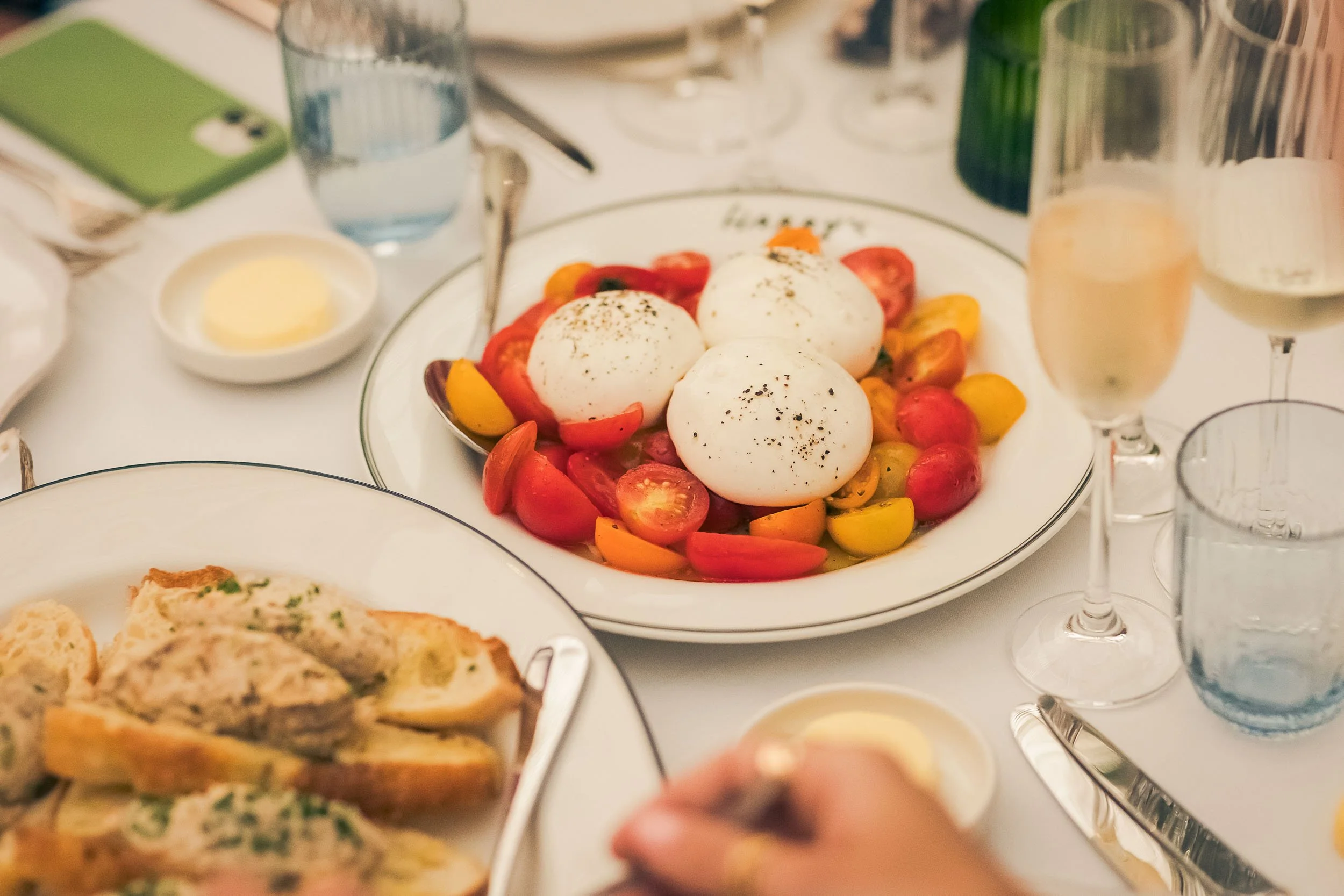 A table set with a plate of colorful cherry tomatoes topped with three scoops of burrata cheese, sprinkled with black pepper, surrounded by glasses of wine and water, and plates of bread and butter.