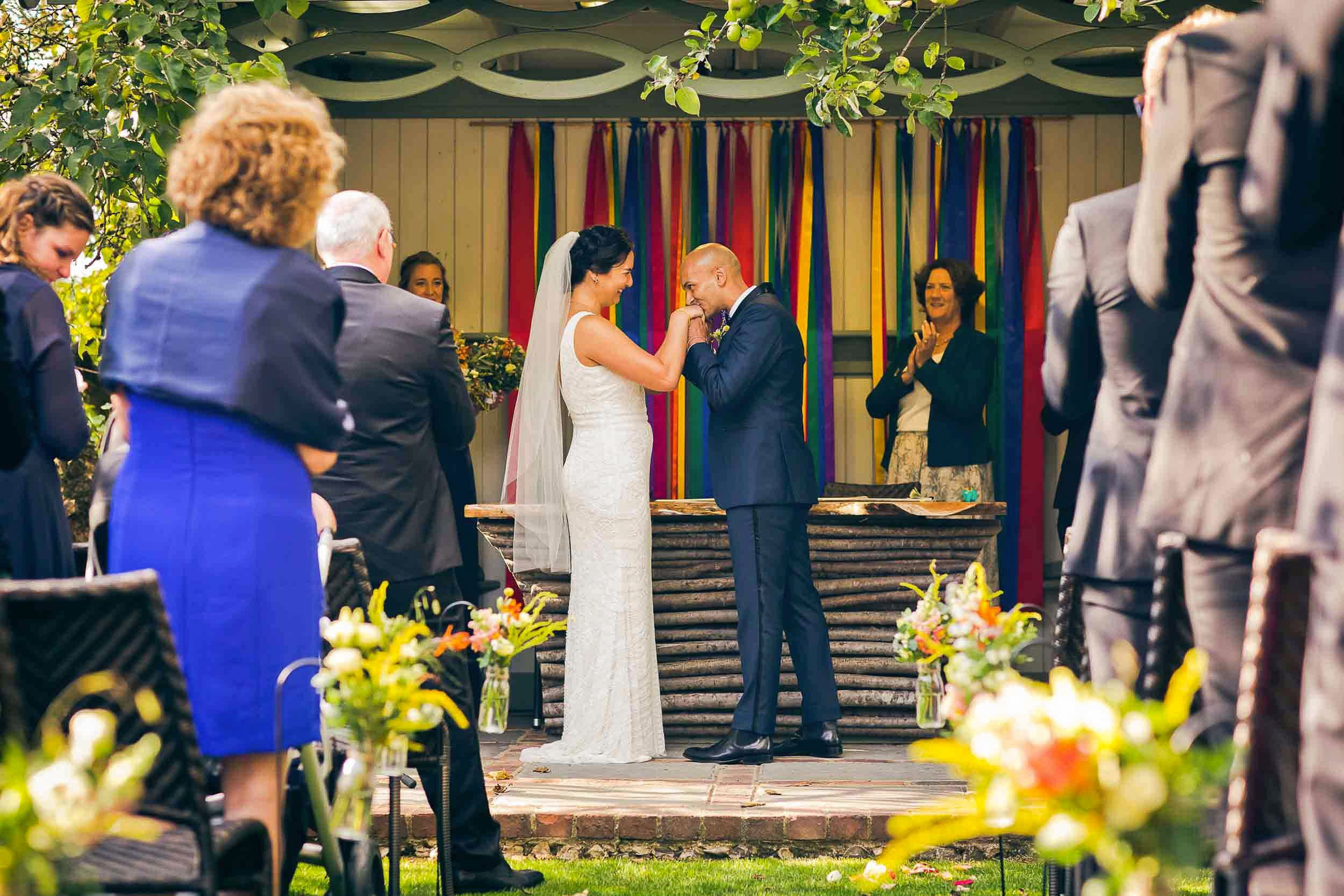 A wedding ceremony outdoors beneath a wooden pavilion decorated with colorful streamers. The bride and groom are in the center, holding hands and smiling, with the groom kissing the bride's hand. Guests are seated on either side, clapping and watchin