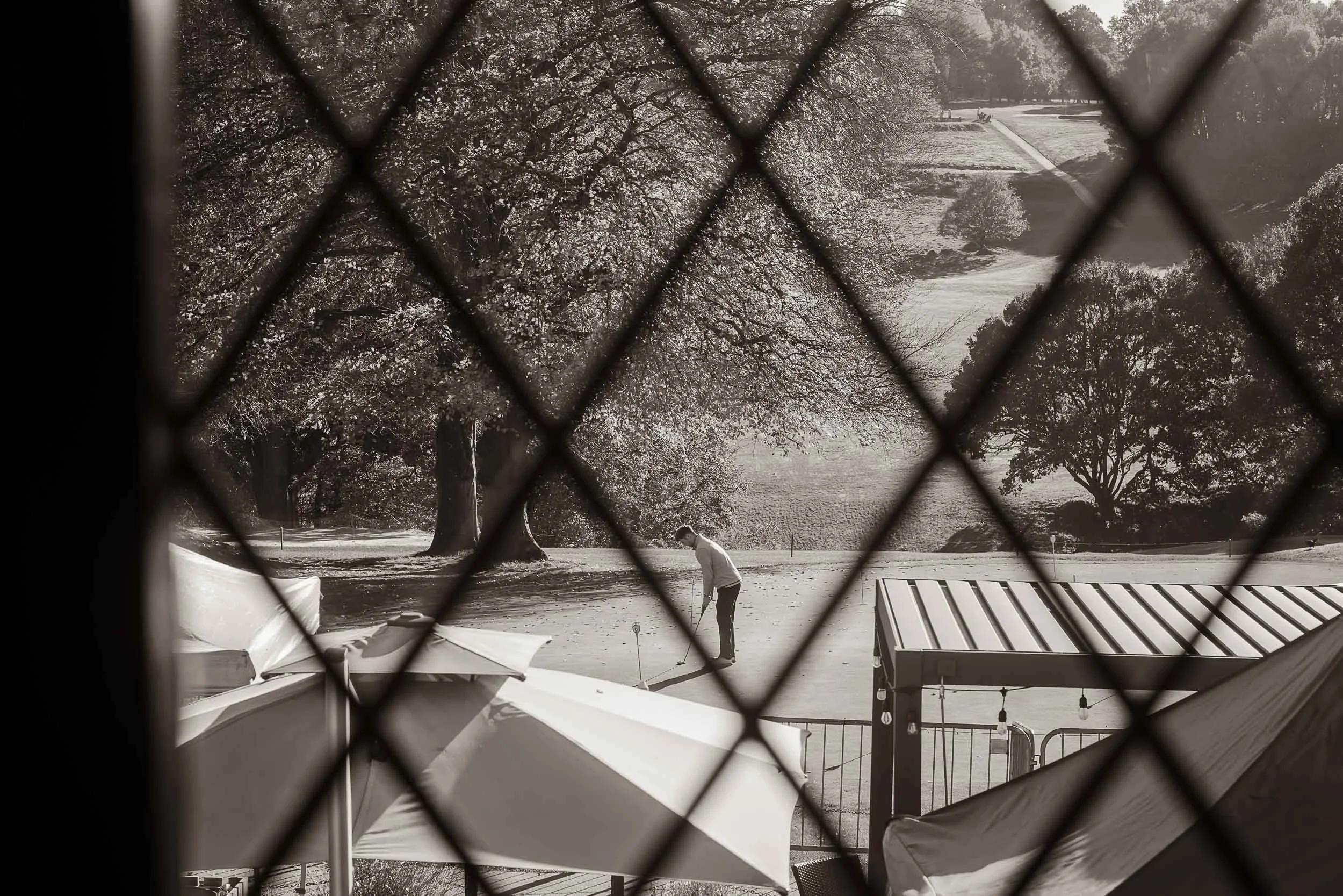 A person playing mini-golf on a course surrounded by trees, viewed through a window with diamond-patterned security bars in black and white.