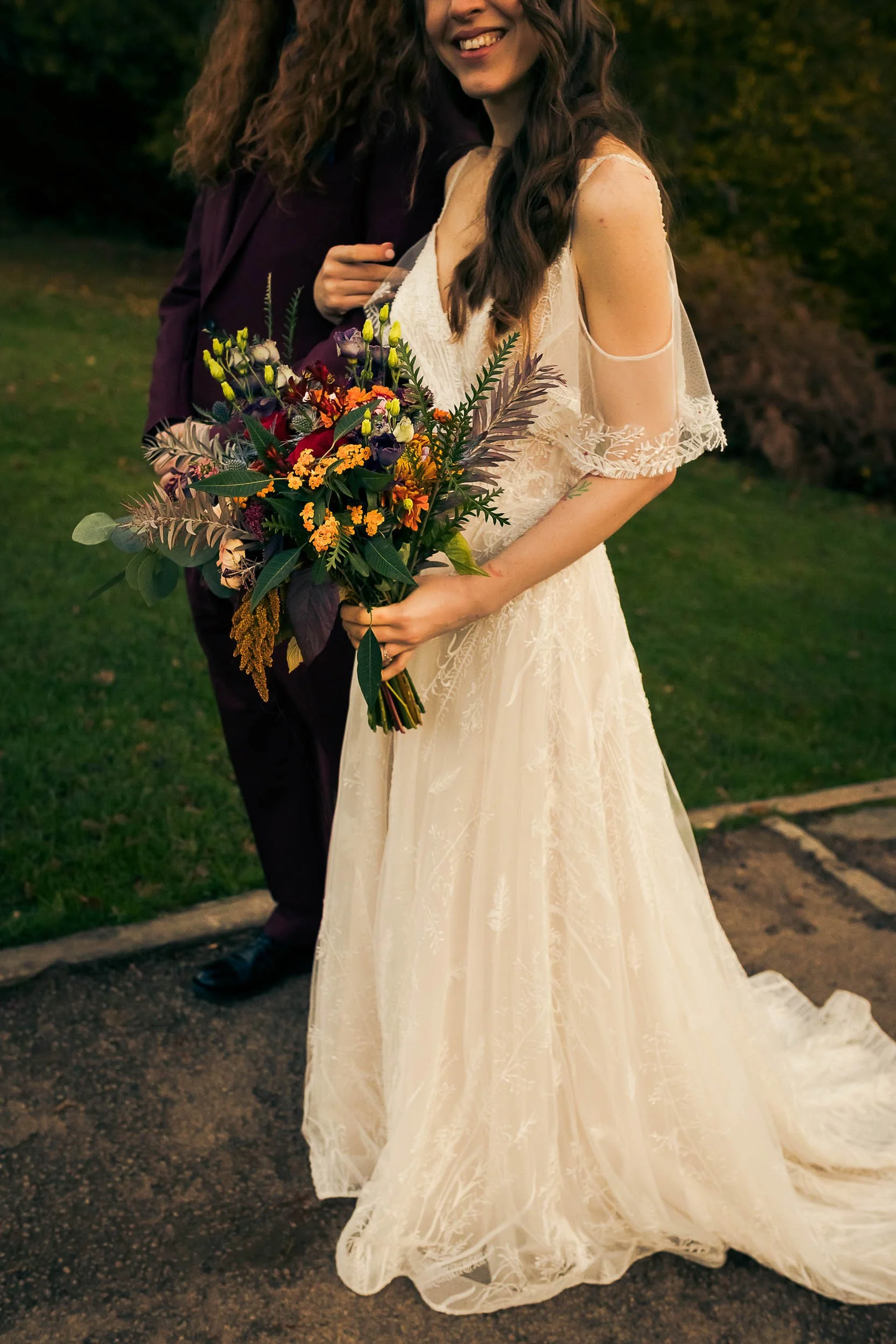 A bride holding a colorful bouquet of flowers outdoors, wearing a lace wedding dress with sheer sleeves, standing on a dirt path.
