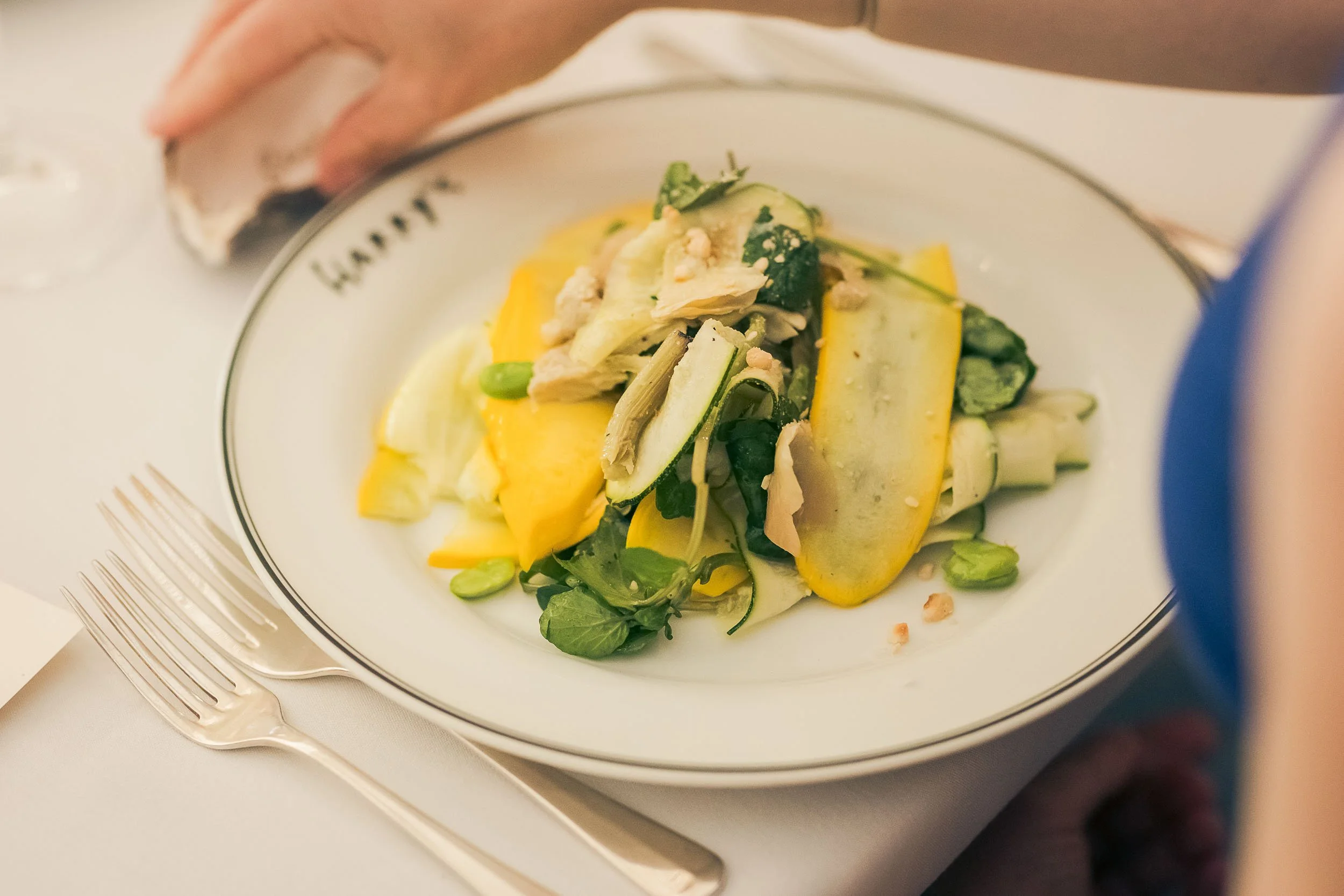 A plate of mixed salad with yellow squash, zucchini, greens, and seeds, next to a fork on a white tablecloth.