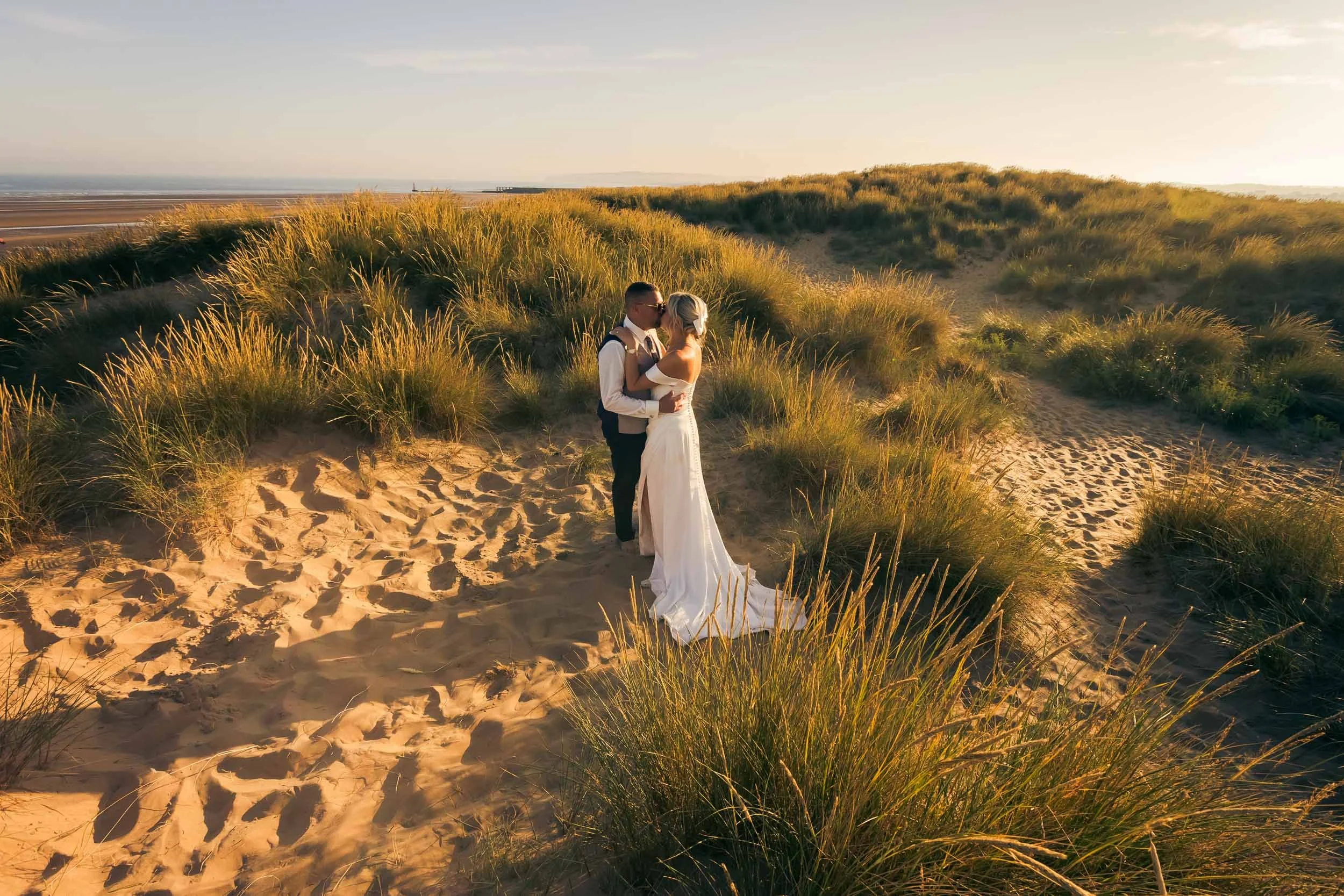A bride and groom standing on sandy beach dunes with tall grasses, embracing during sunset.