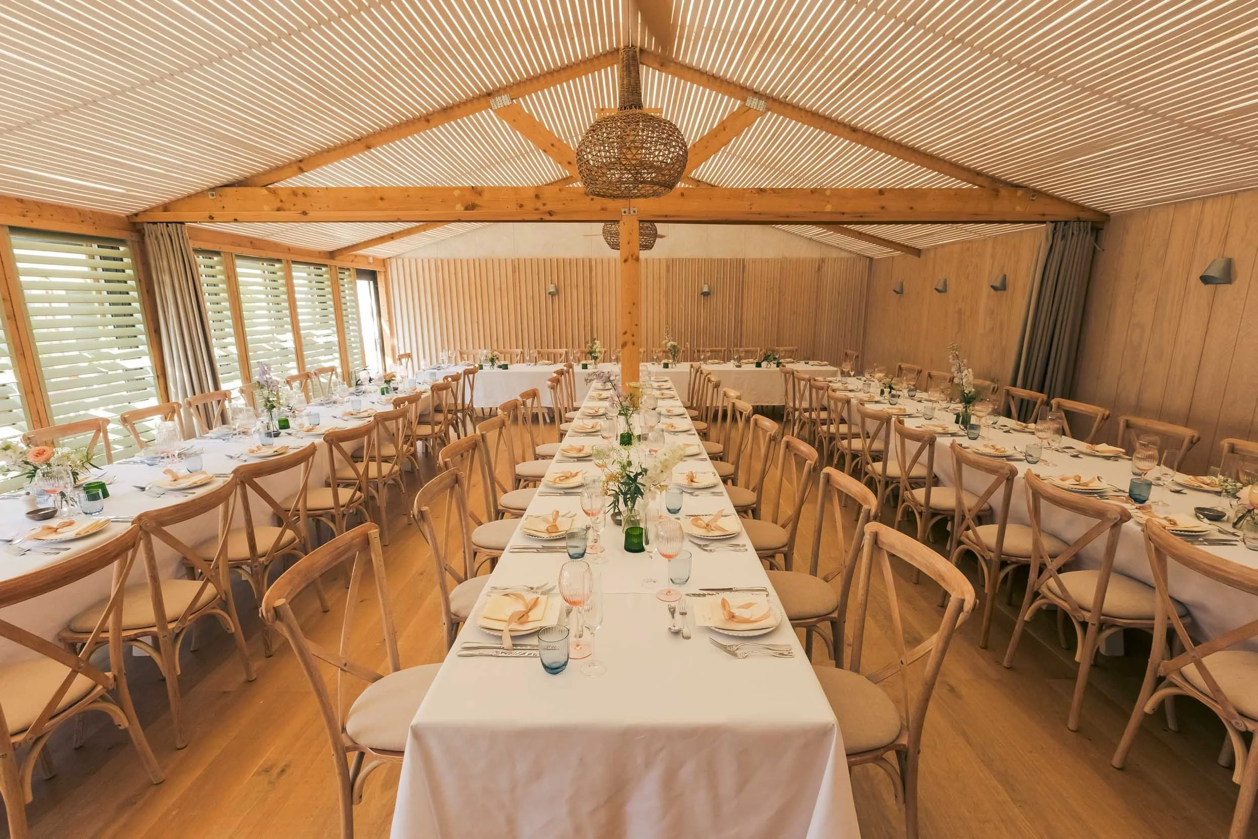 Interior of a wood-paneled dining room arranged for a formal event with multiple long tables, white tablecloths, chairs, floral centerpieces, and place settings, natural light coming through windows with blinds.