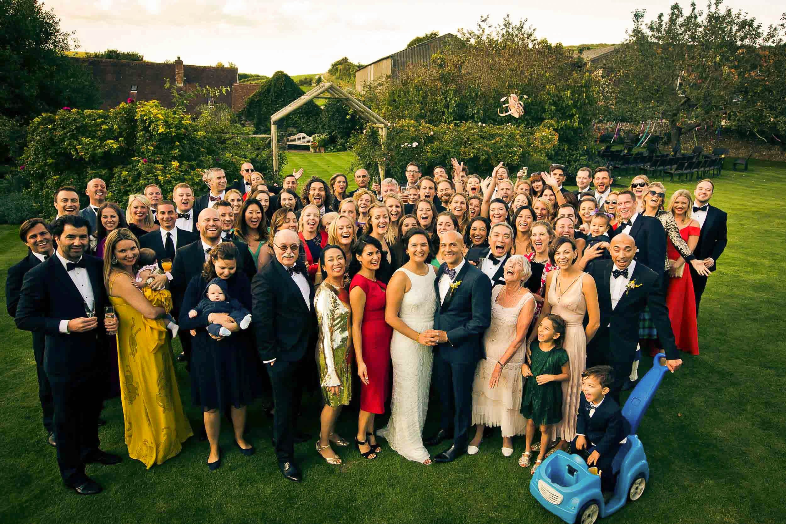 A large group of people dressed in formal attire gathered outdoors in a garden for a celebration, with some children present and a man riding a blue toy car.