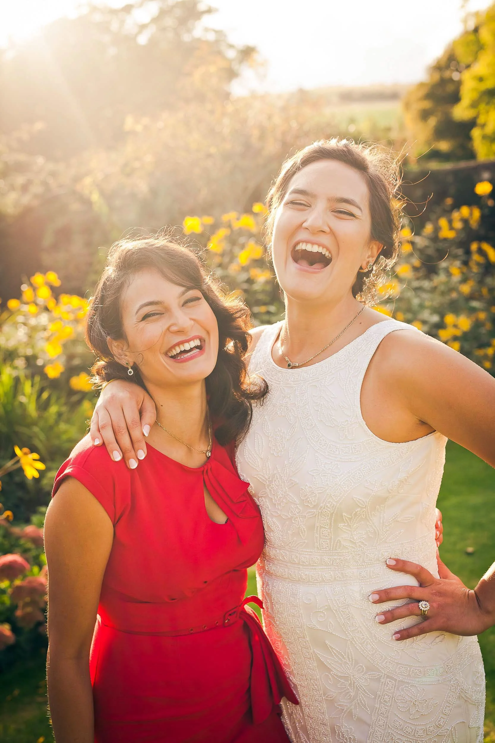 Two women smiling and laughing outdoors during sunset, standing close together with one woman's arm around the other's shoulder. One woman is wearing a red dress, and the other is wearing a white lace dress.