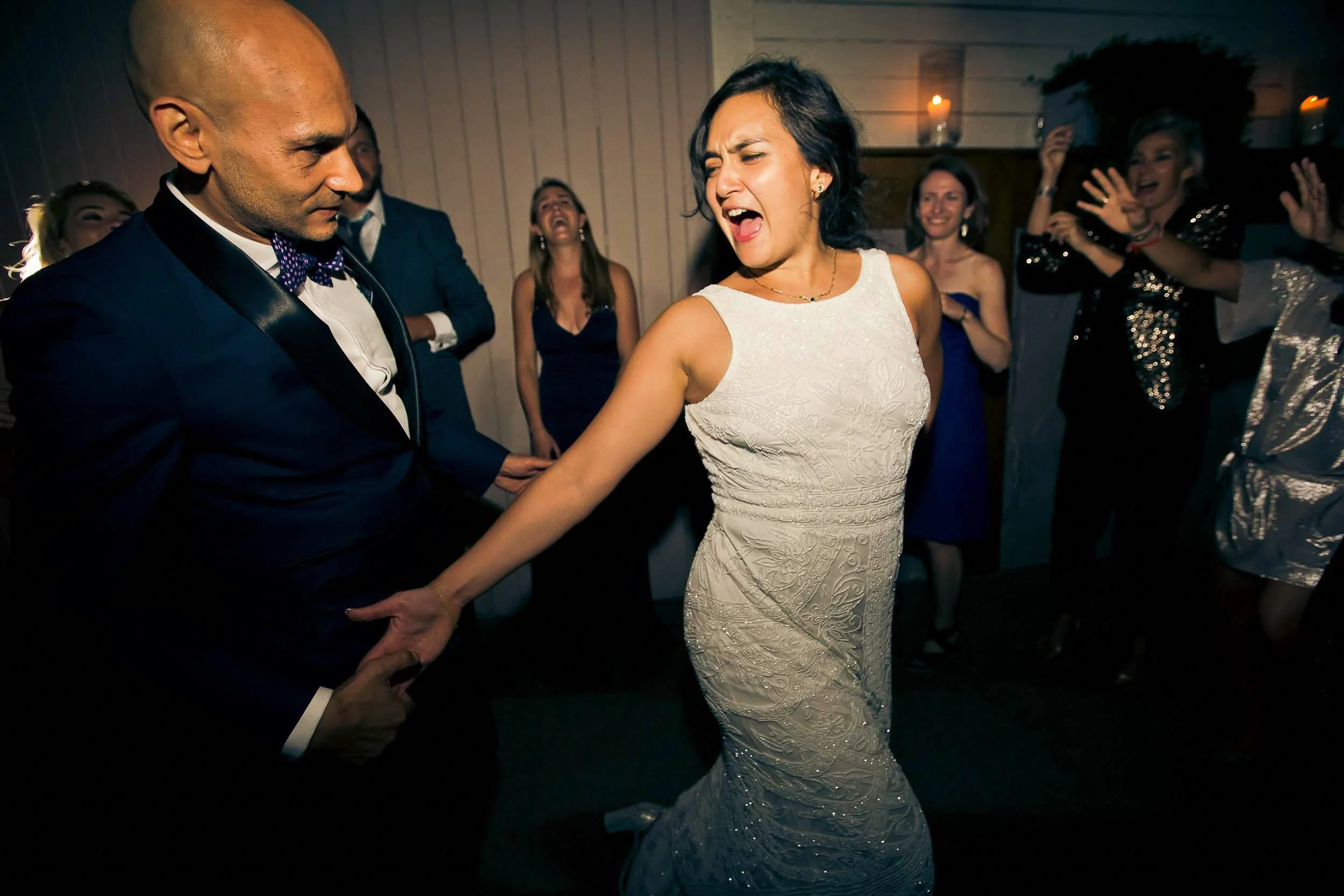People dancing at a celebration or party, with a woman in a white dress dancing and a man in a tuxedo nearby, surrounded by others enjoying the moment.