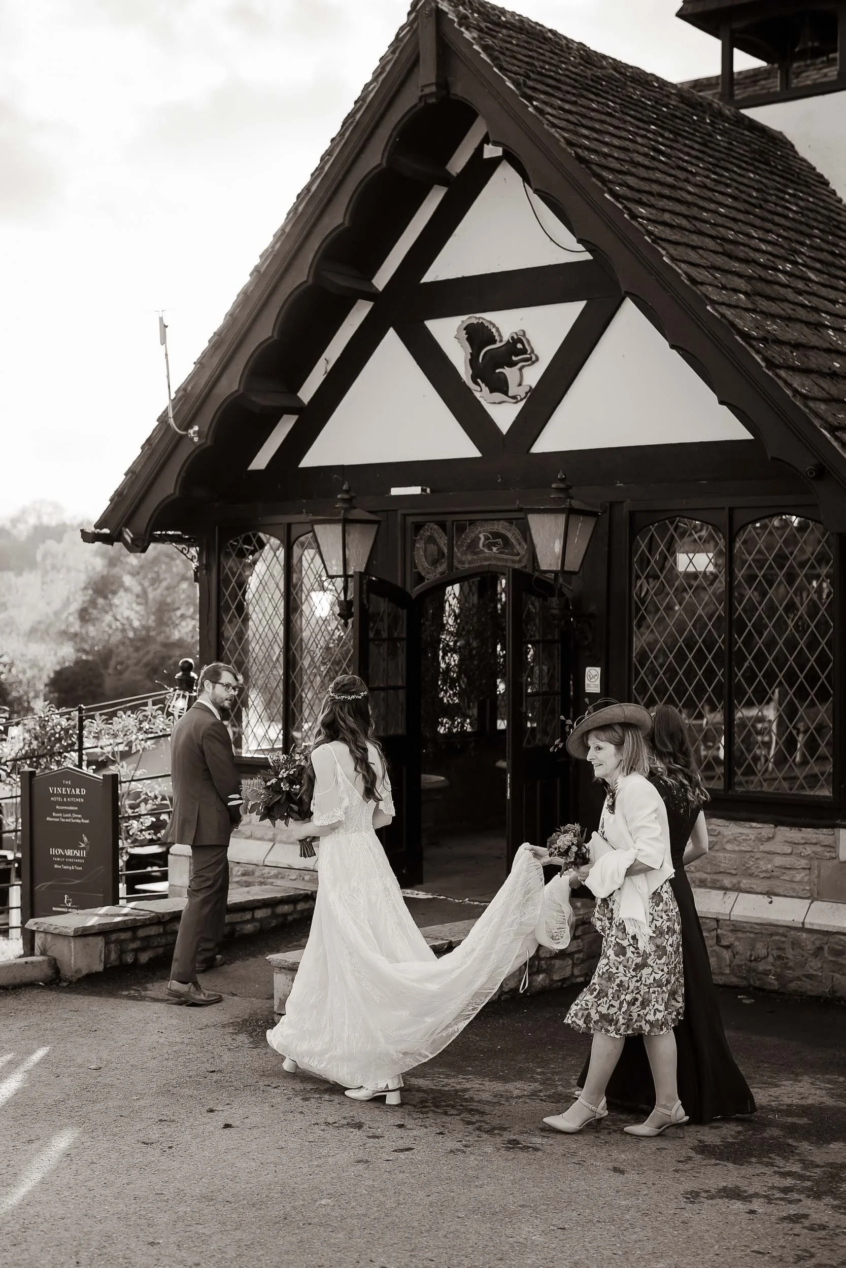 Wedding ceremony outside a building with Gothic architecture, featuring a bride in a long white dress holding flowers, accompanied by two women, and a man standing nearby.