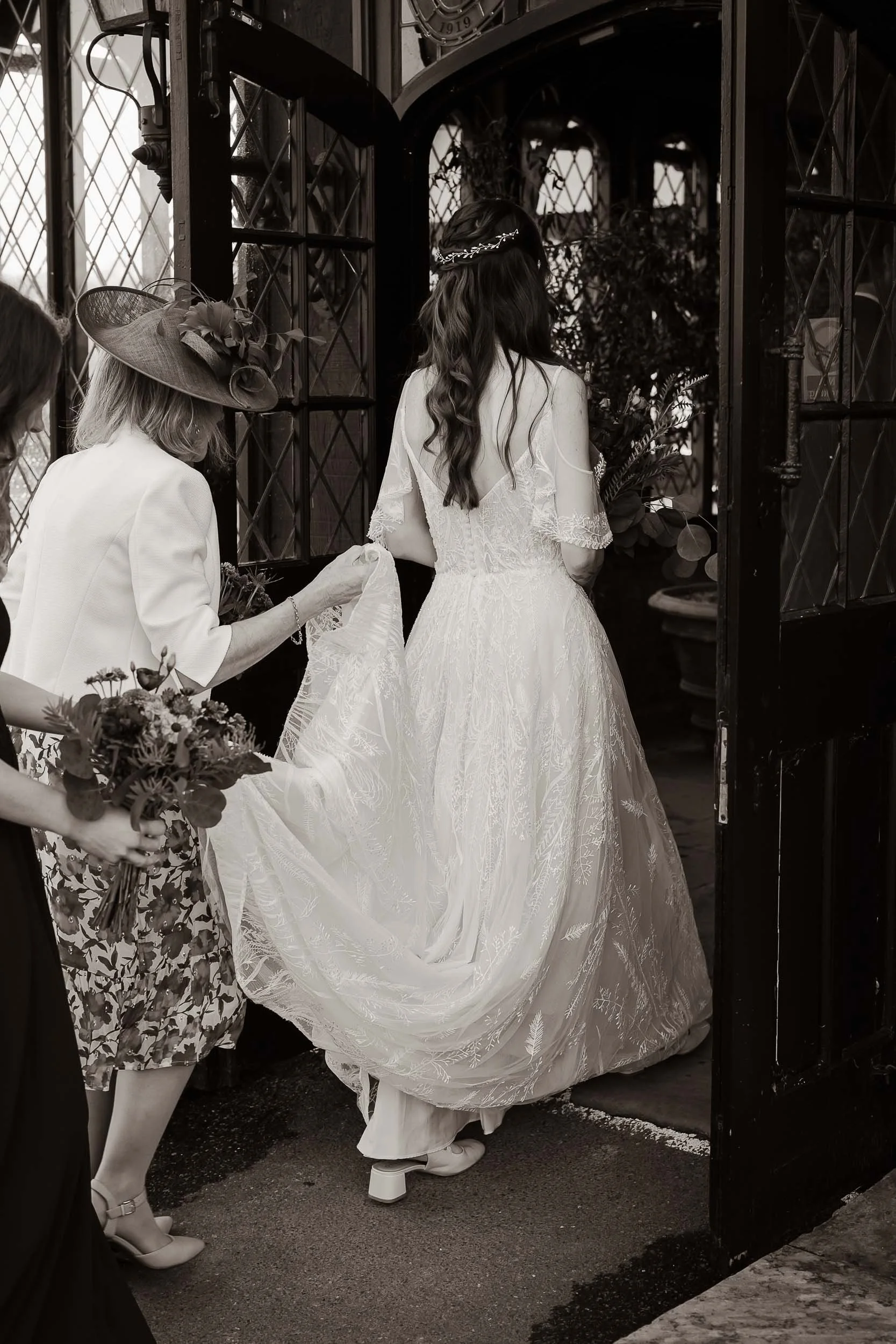 A bride in a vintage lace wedding dress with a deep back, walking through a dark doorway, helped by women including one in a large hat, during her wedding ceremony.