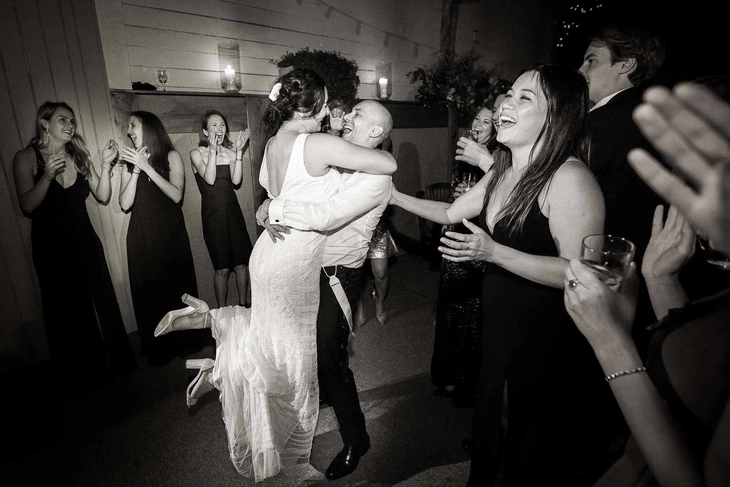 People celebrating at a wedding reception, with a couple dancing and surrounded by friends who are clapping and smiling.
