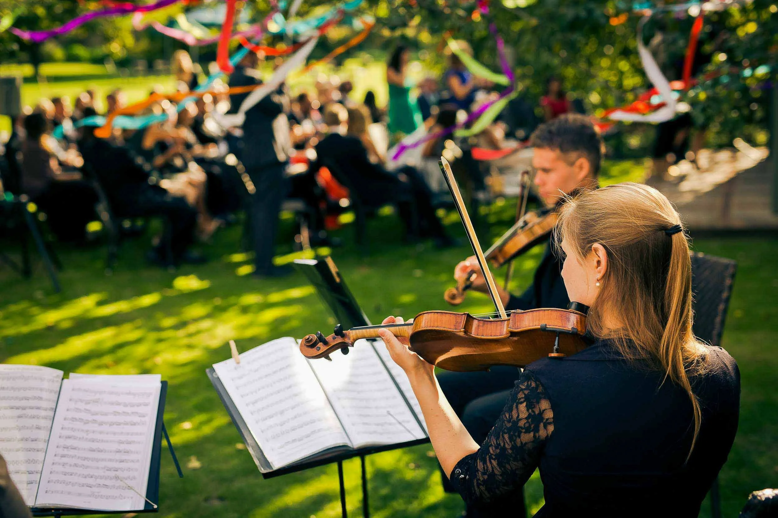 Group of musicians, including a woman playing the violin, performing outdoors at a concert in a park with an audience in the background and colorful decorations hanging overhead.