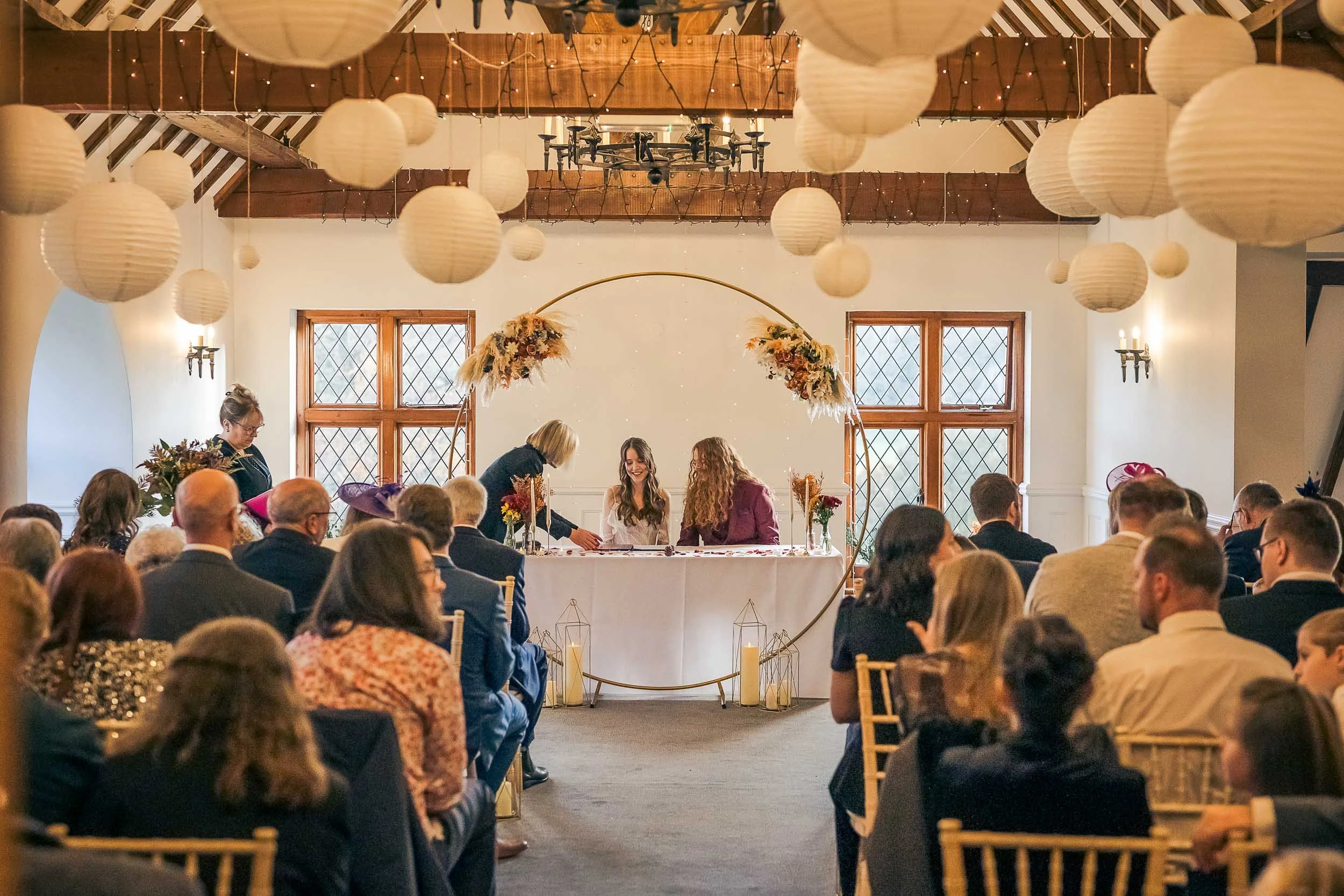 A wedding reception inside a decorated venue with hanging paper lanterns, a circular floral arch, and guests seated at tables facing the bride and groom who are seated at a head table.
