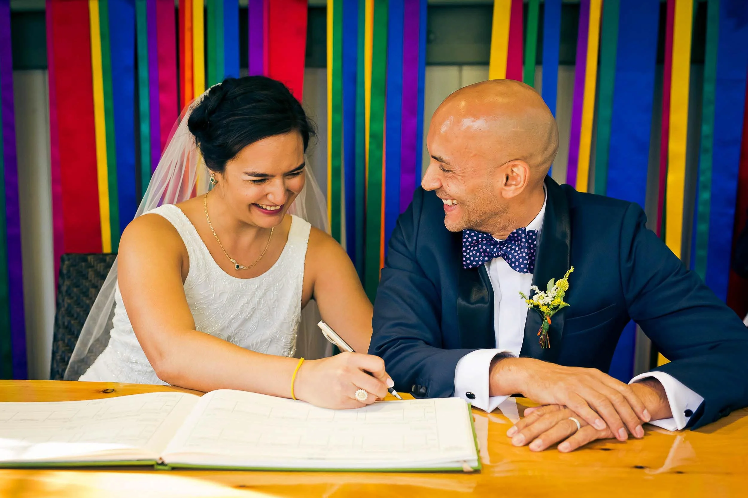 Bride and groom signing marriage register, smiling and laughing, with colorful background.