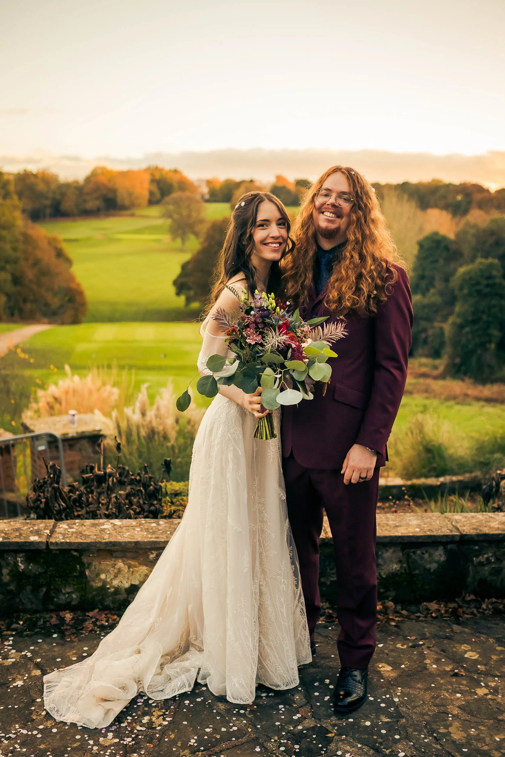A happy couple in wedding attire posing outdoors on a stone patio, with the bride holding a bouquet and a scenic landscape with trees and a golf course in the background during sunset.