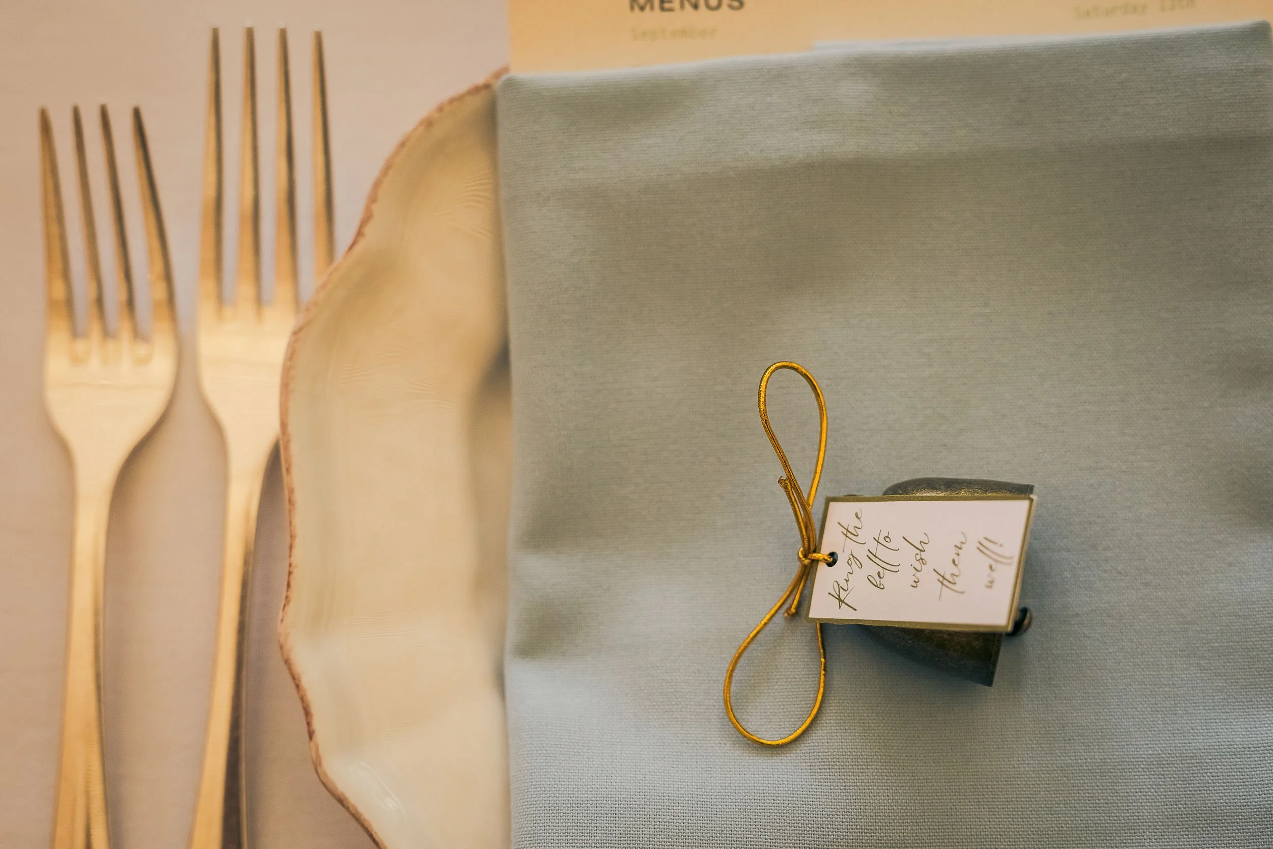 Table setting with two gold forks, a seashell, a light blue napkin with a small black box gift, and a note attached with a gold string that reads "Plant the little wilder."
