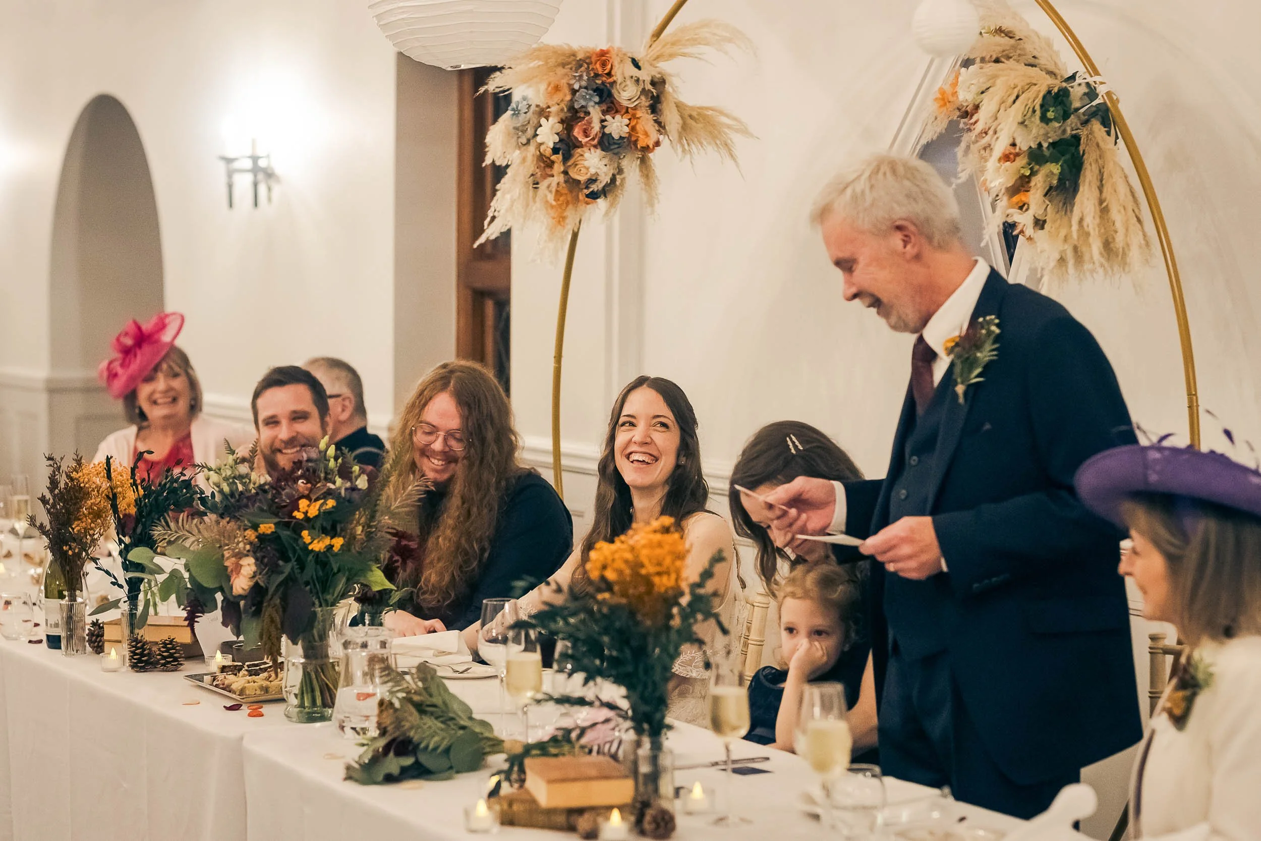 Group of people celebrating at a wedding reception, with a man giving a speech, seated at a decorated table with flowers and champagne glasses, in a warmly lit room with floral and pampas grass decorations.