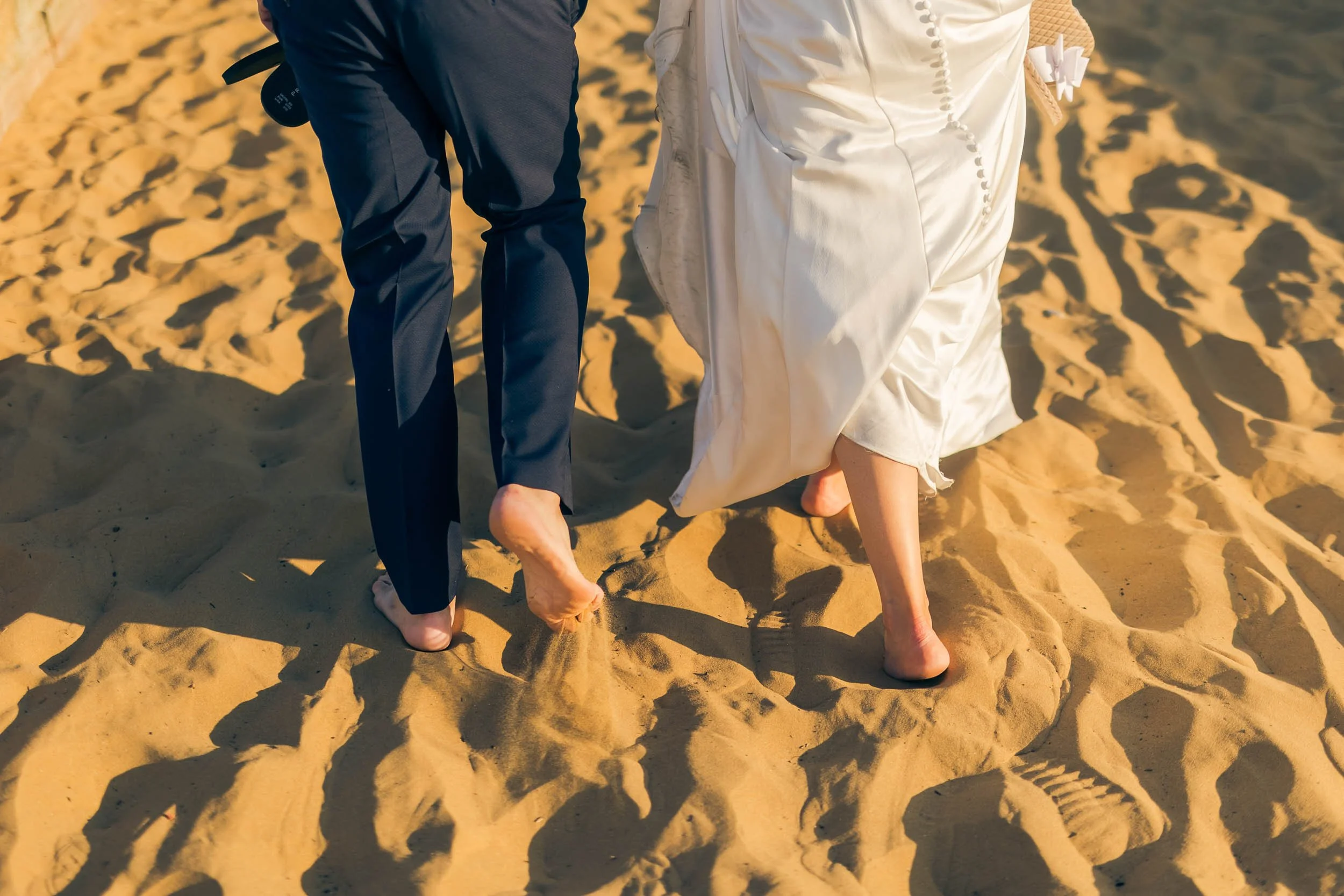 A couple walking barefoot on a sandy beach, with only their legs and feet visible, dressed in formal wedding attire.