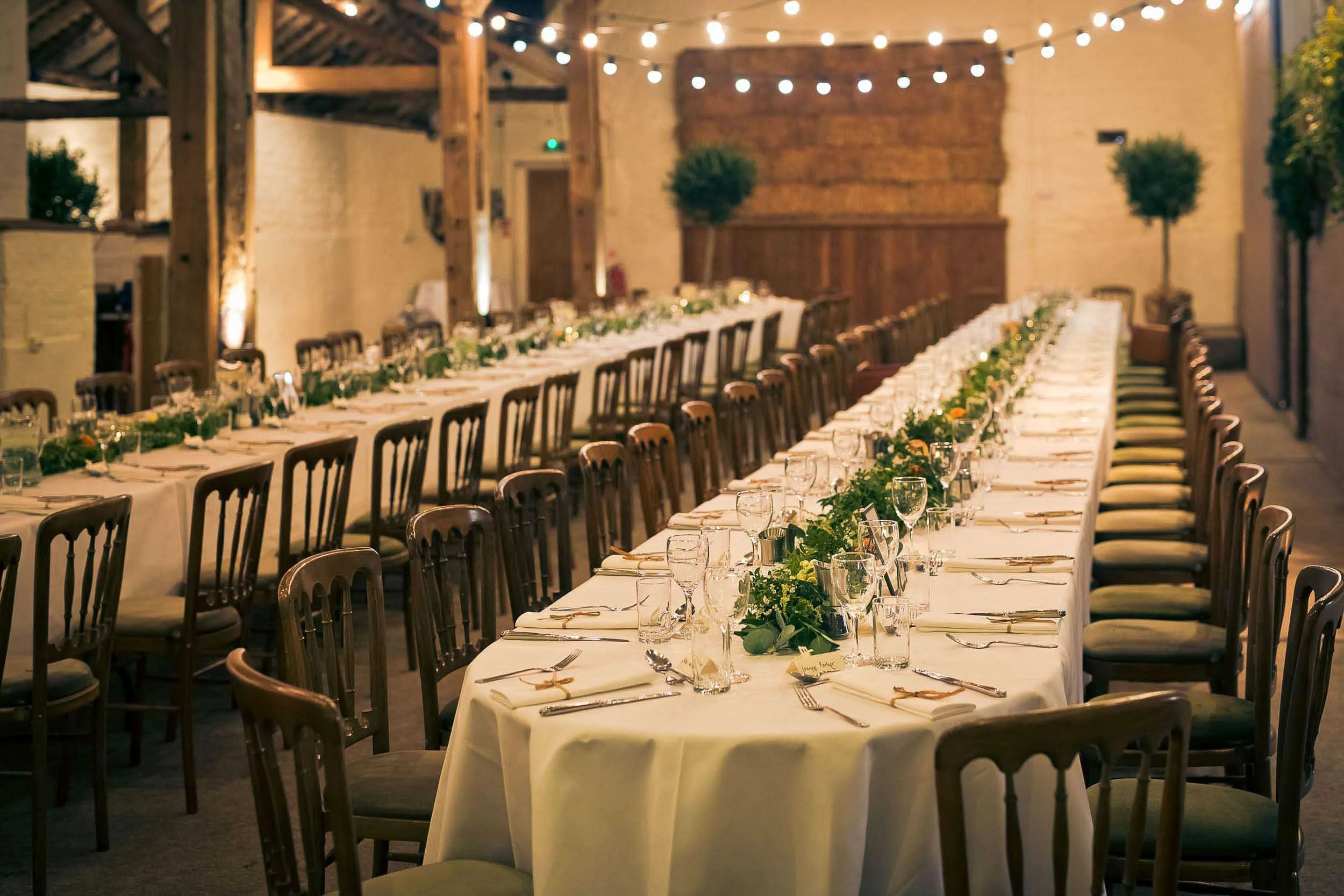 Long banquet table set for a formal event with white tablecloths, glassware, plates, silverware, and green foliage centerpieces in a rustic hall with exposed wooden beams and string lights.