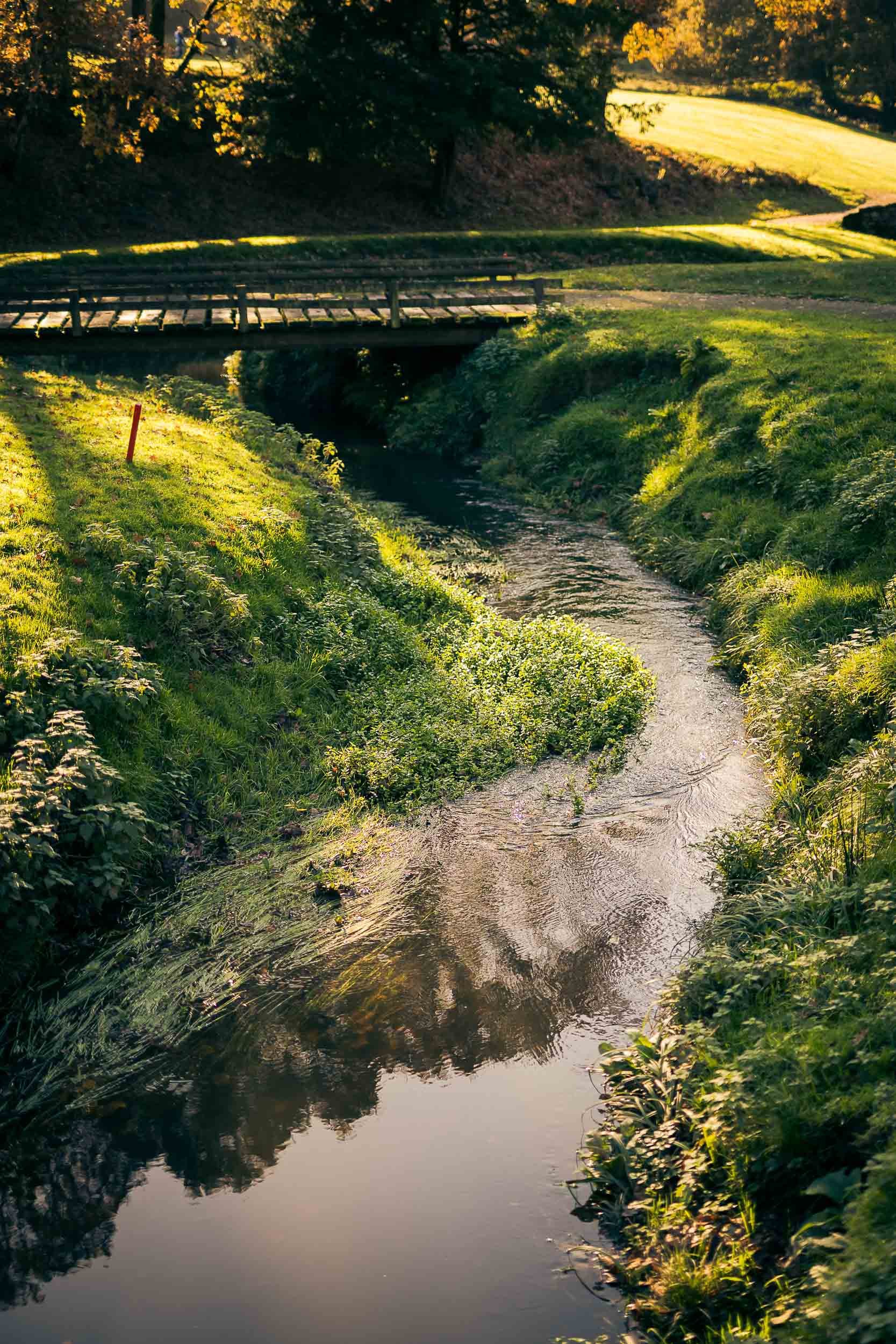 A small stream flowing through lush green grass with a wooden bridge in the background, surrounded by trees and sunlight.