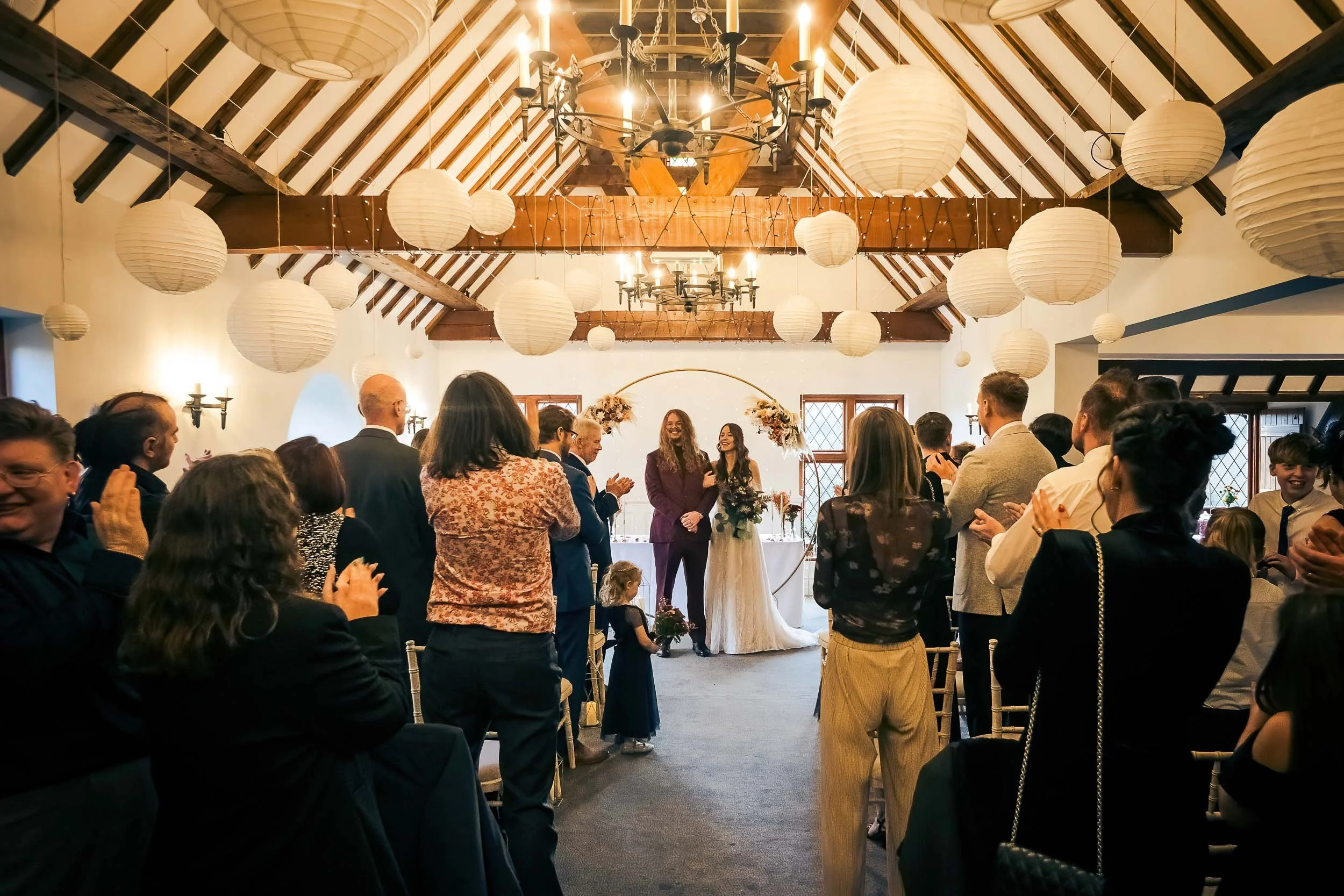 A wedding ceremony inside a decorated venue with guests clapping for the couple, who are standing at the altar. The ceiling has hanging lanterns and a chandelier, with a floral arch behind the couple.