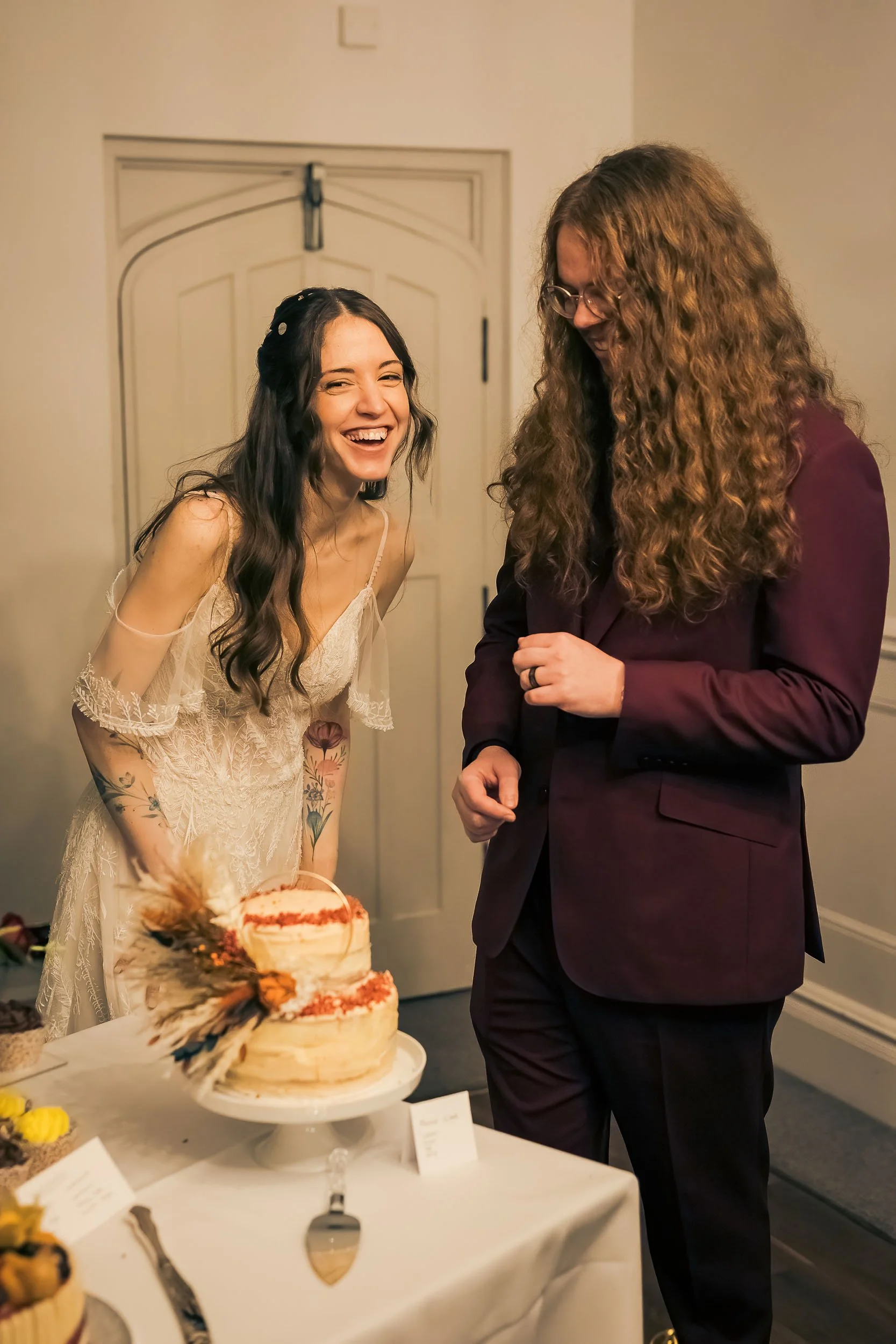 A joyful bride with long dark hair and tattoos on her arms is laughing, beside a woman with long curly hair in a maroon suit, at a wedding reception with a cake on the table.