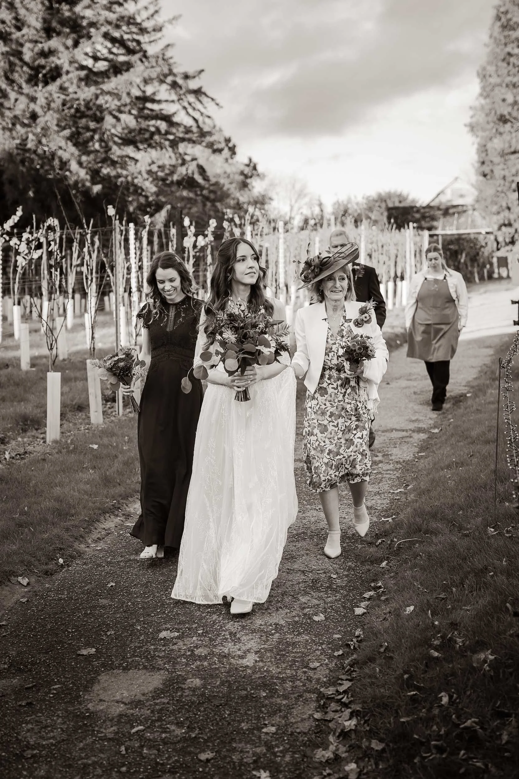A group of women walking on a garden path, holding bouquets, dressed in formal and semi-formal attire. One woman is wearing a white dress and hat, possibly a bride, with others in darker dresses.